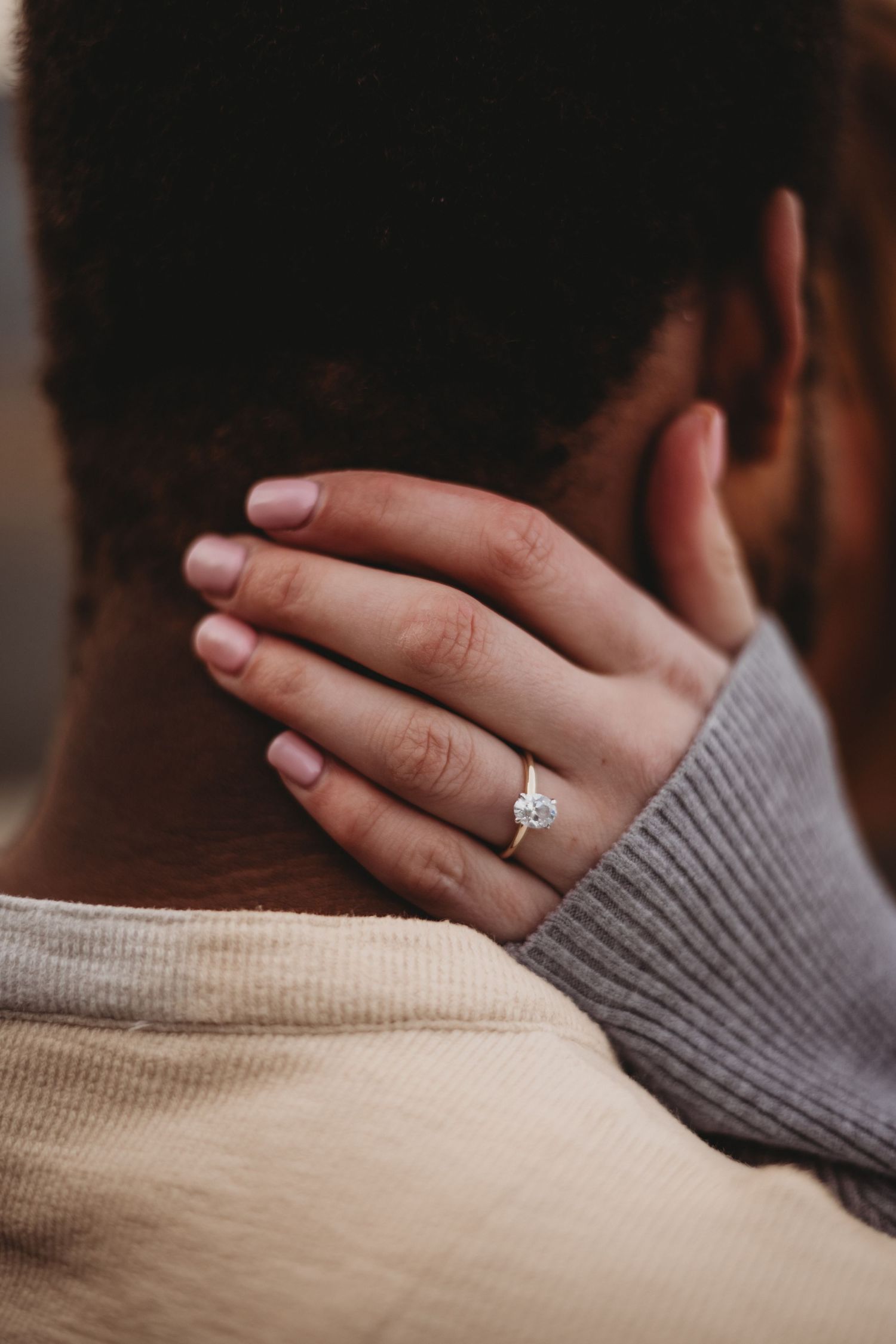 Close-up of a woman's hand with a solitaire diamond engagement ring resting on her partner's neck.