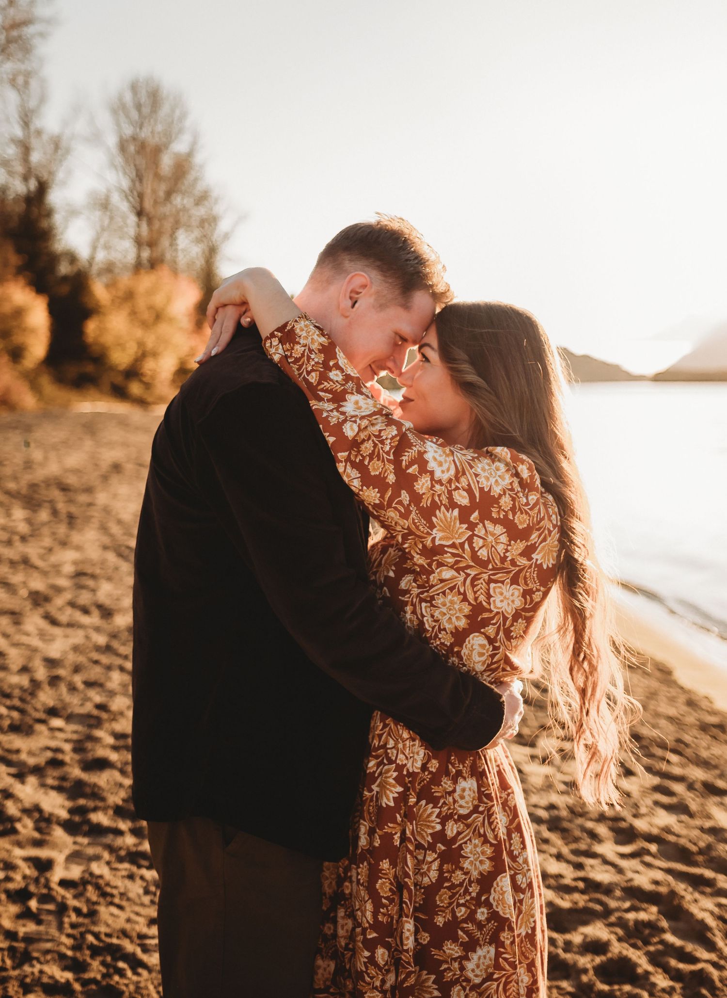 Couple in golden sunset light touching foreheads and embracing on a sandy lakeshore beach.
