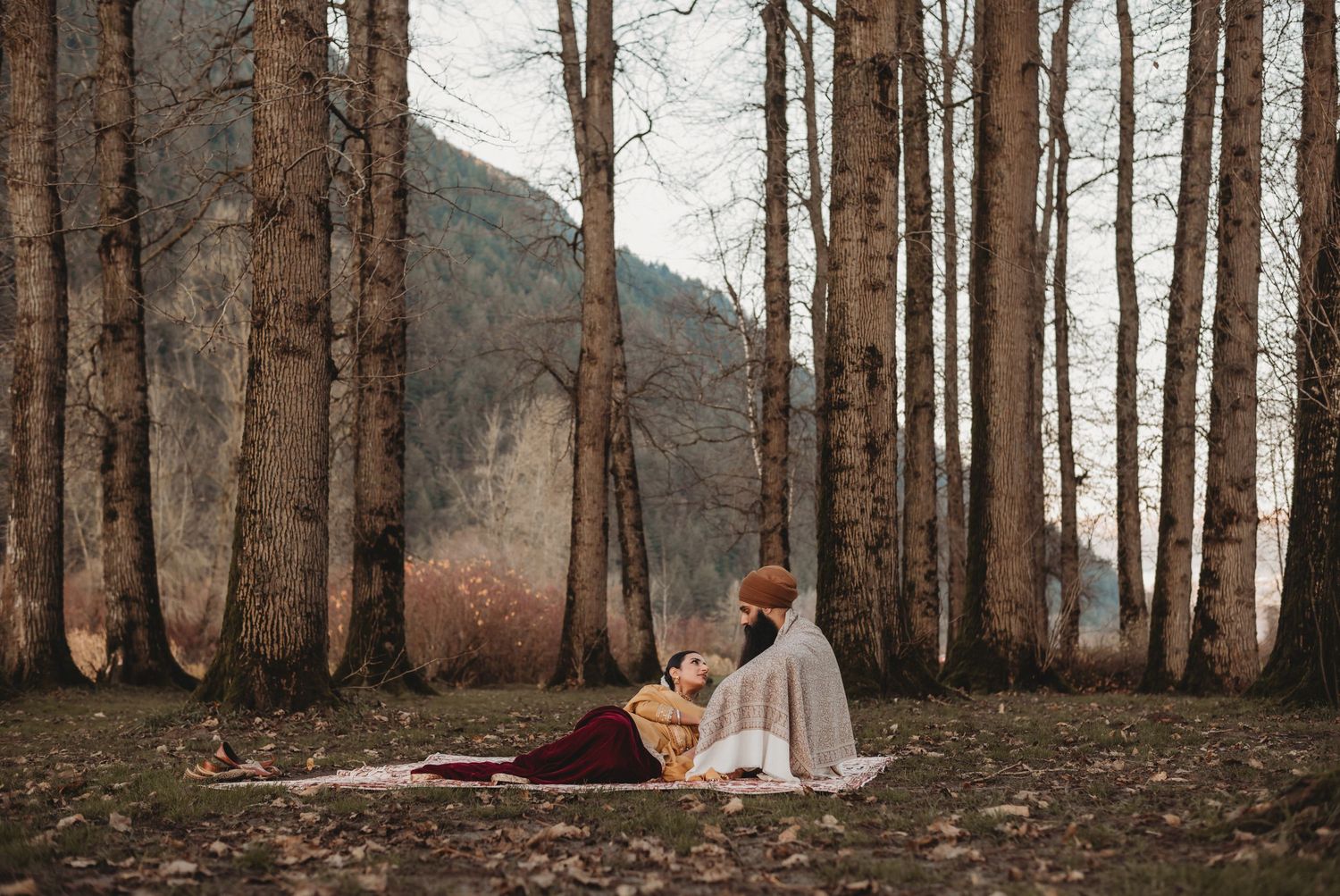 Sikh couple sitting together on a rug in a forest, surrounded by tall trees and autumn foliage.
