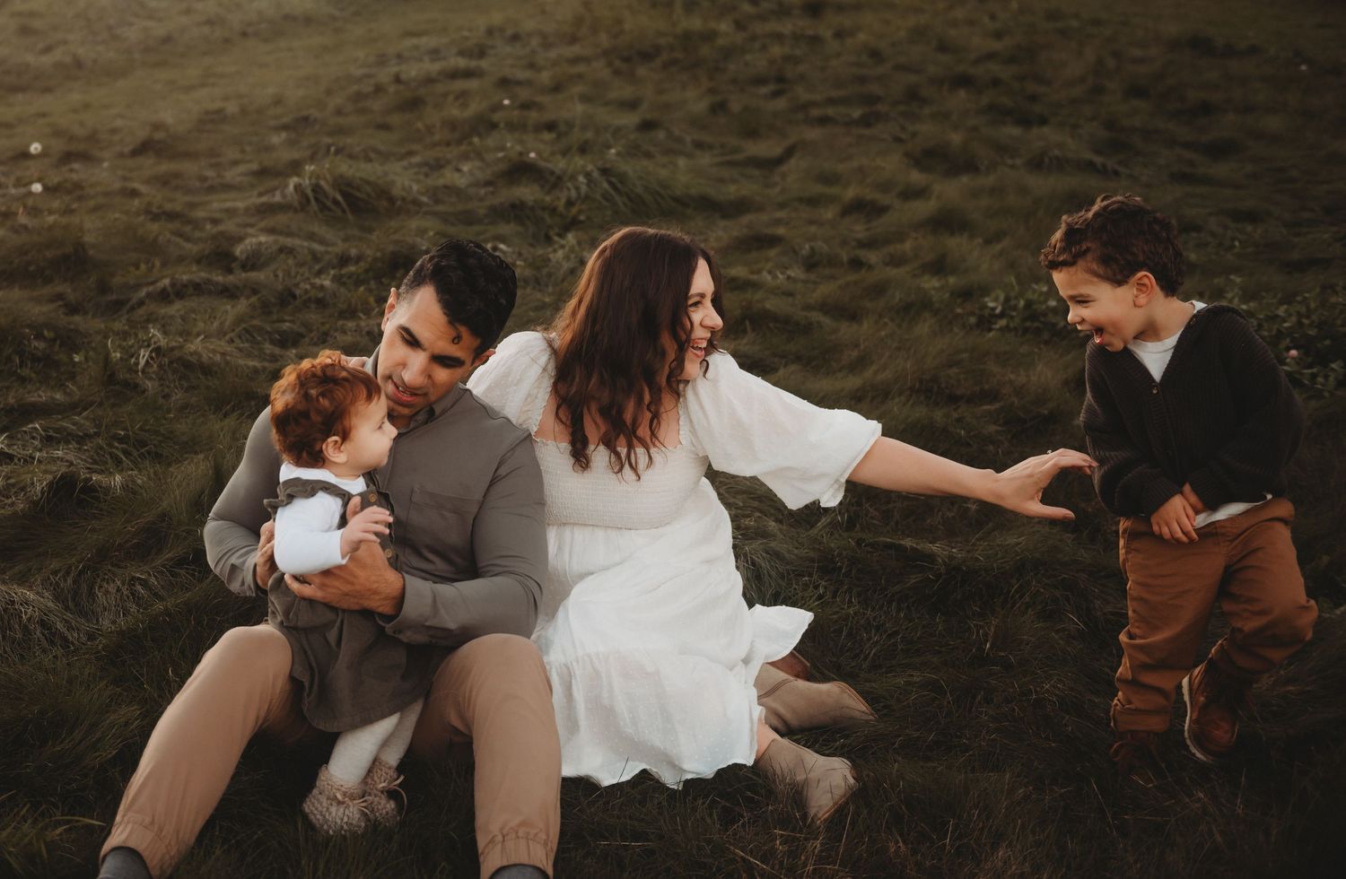 A joyful family laughs and plays together on a grassy field, with two young children and their parents.