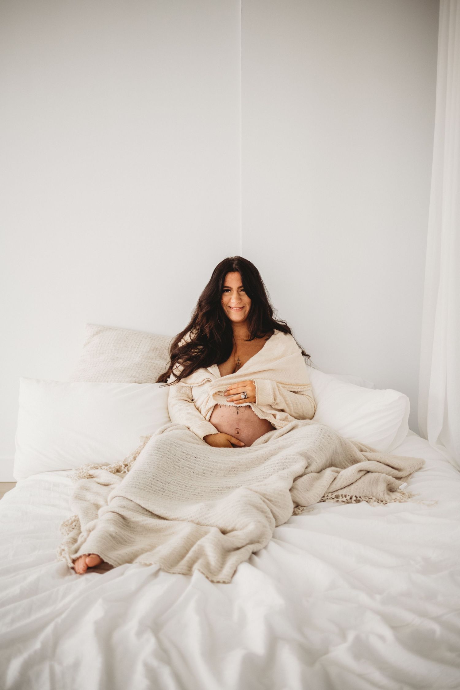 Smiling pregnant woman wrapped in cream blanket sitting on white bed.