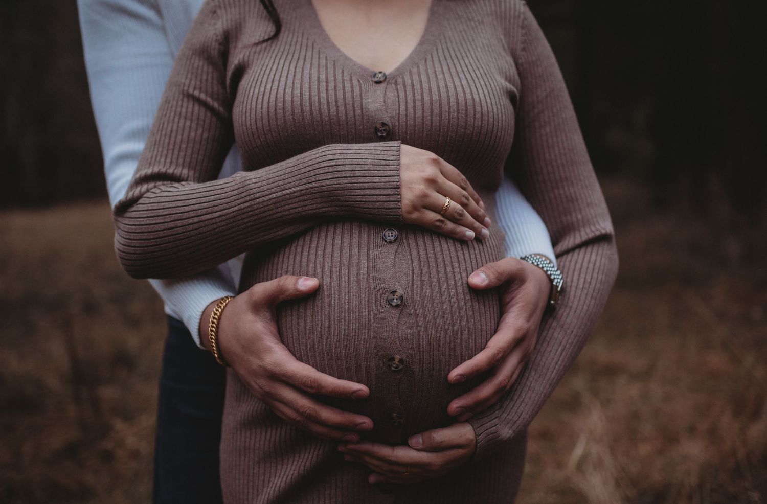Couple cradling pregnant belly outdoors, close-up of hands on baby bump in forest setting.