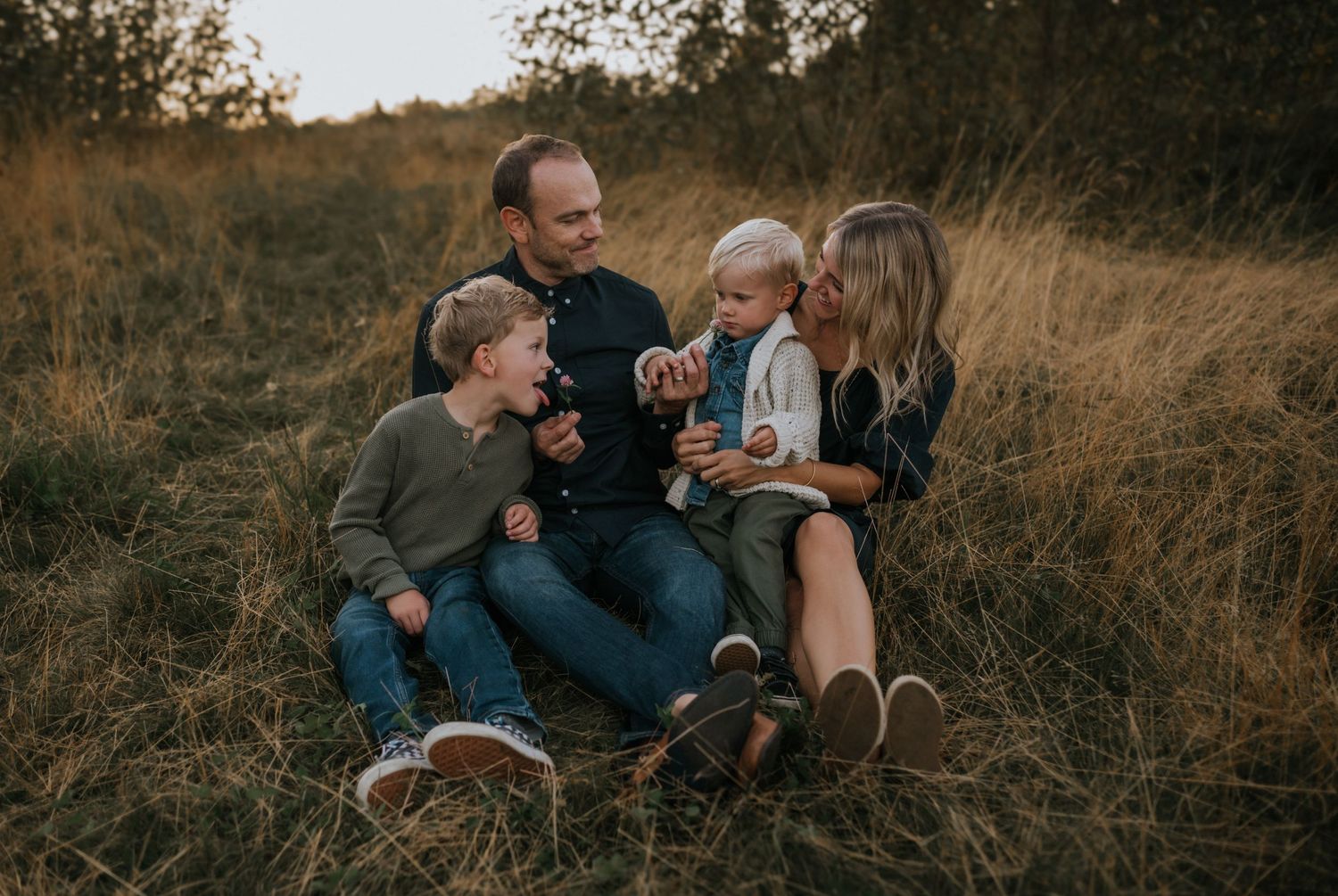 Happy family of four sitting in tall autumn grass laughing together during golden hour.