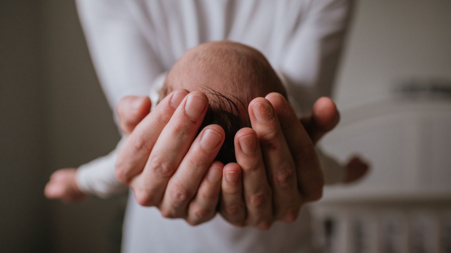 Father gently cradling tiny newborn baby head in both hands in soft natural light.
