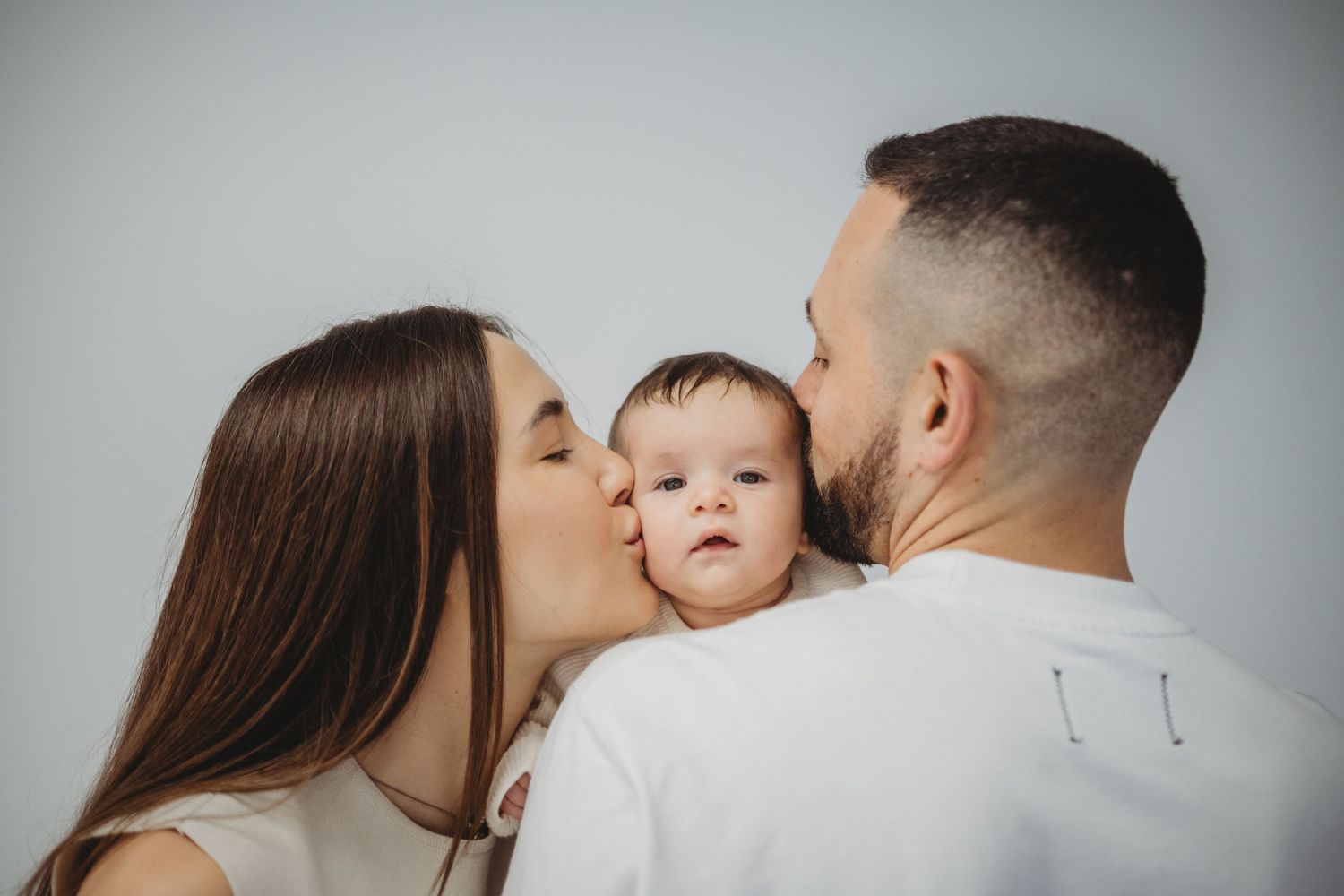 Loving parents kissing their baby on both cheeks against white background.