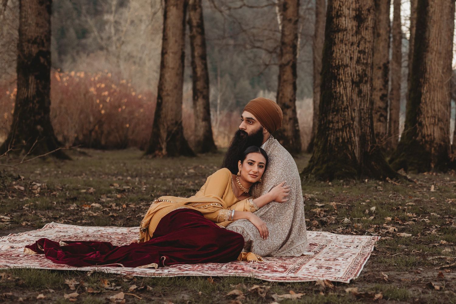 Sikh groom embracing his bride as she leans against him on a rug in an autumn forest setting.