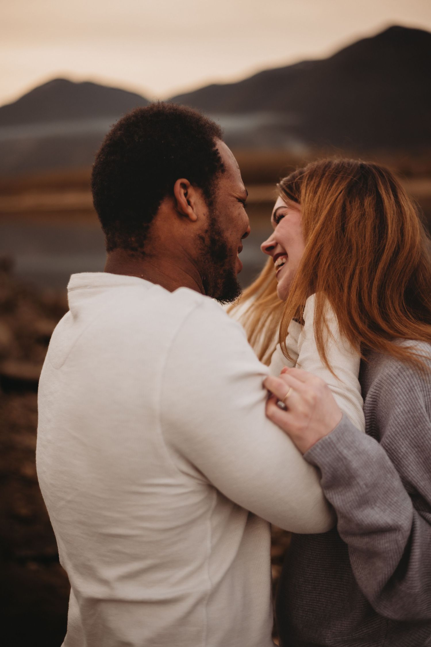 Joyful interracial couple laughing and embracing outdoors with mountains in the background at sunset.