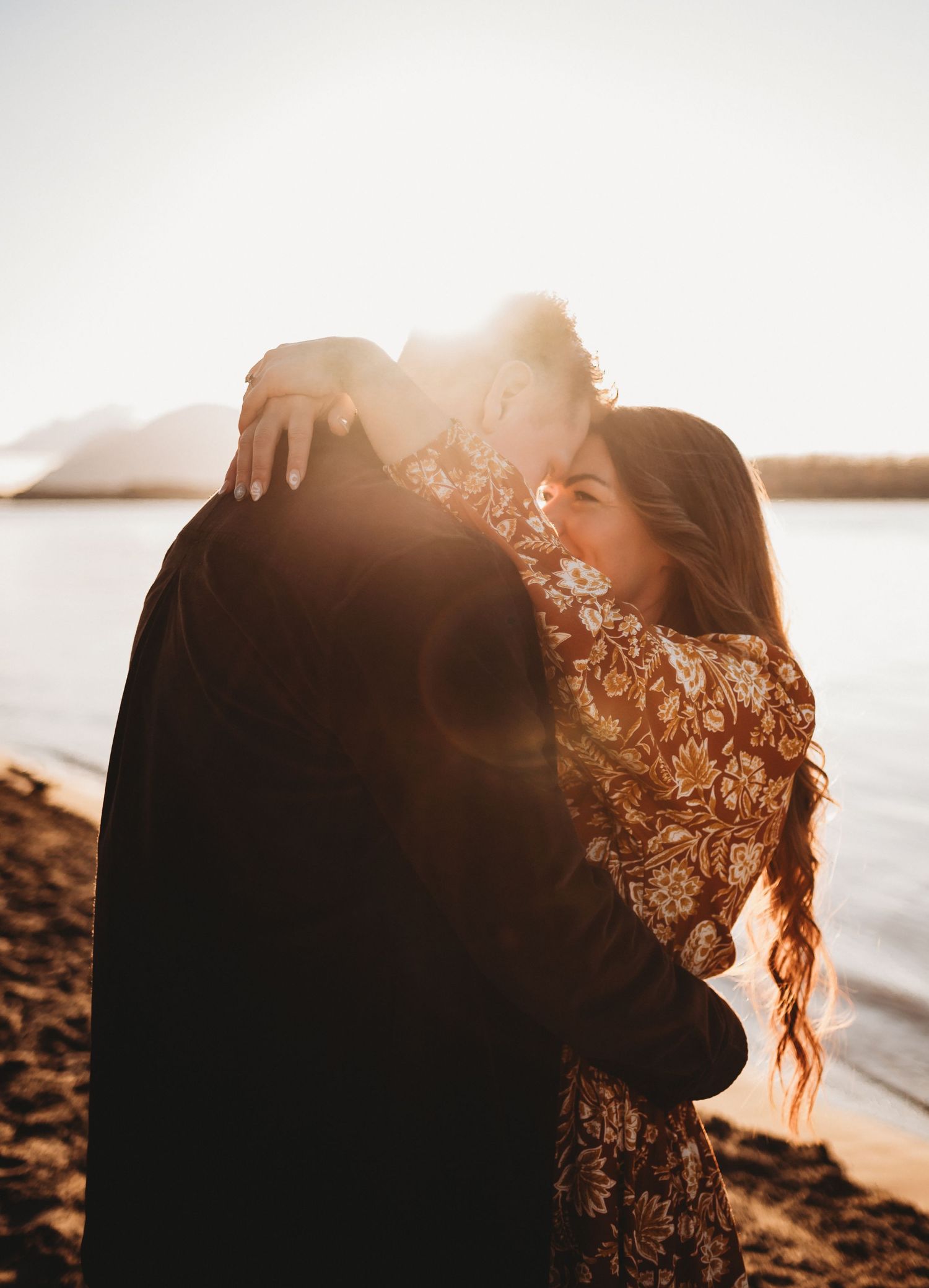 Couple sharing a warm embrace on a sunny beach with golden lens flare and lake backdrop.
