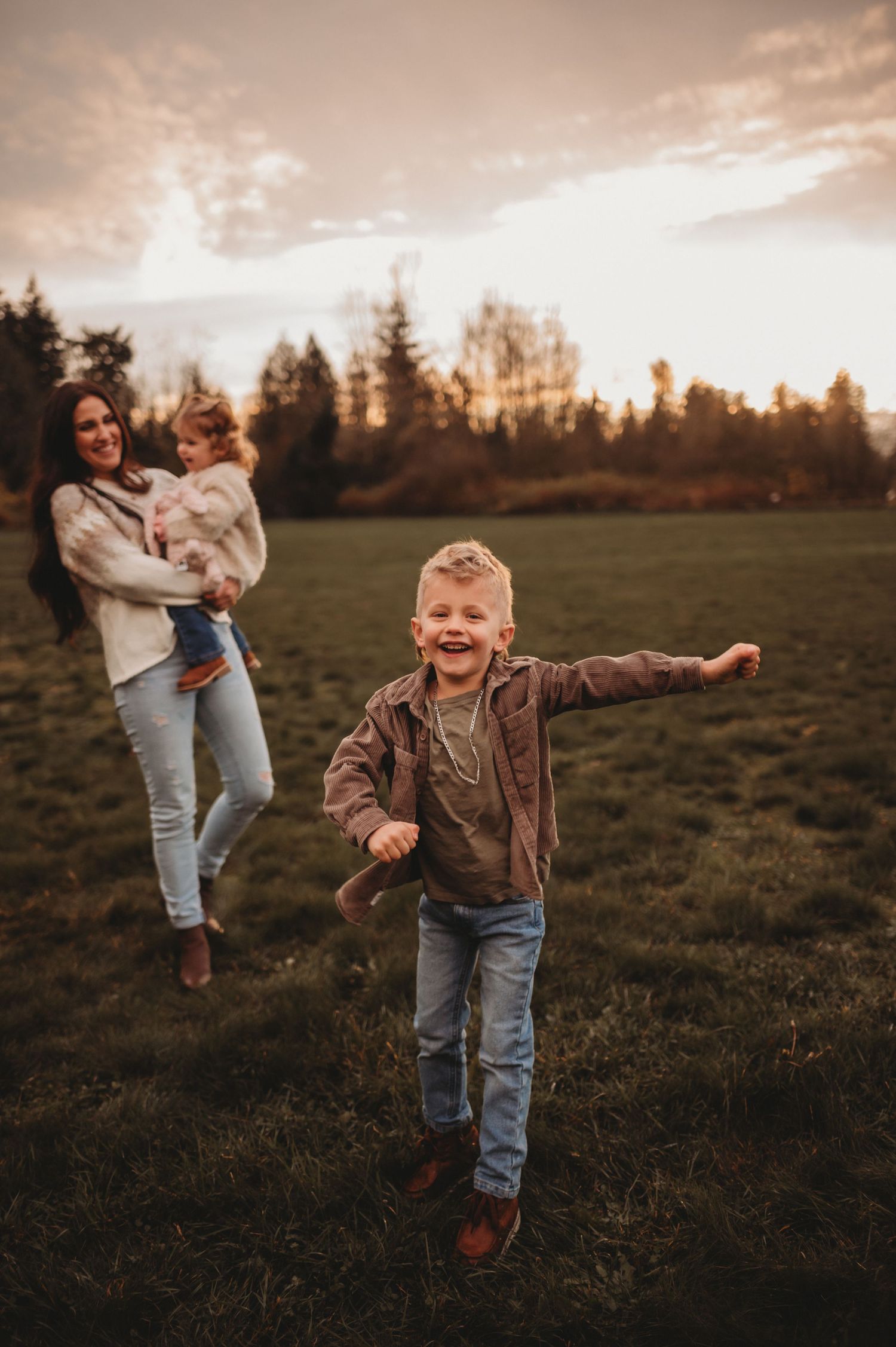 A joyful boy runs toward the camera as his mother holds a toddler in a sunset field.