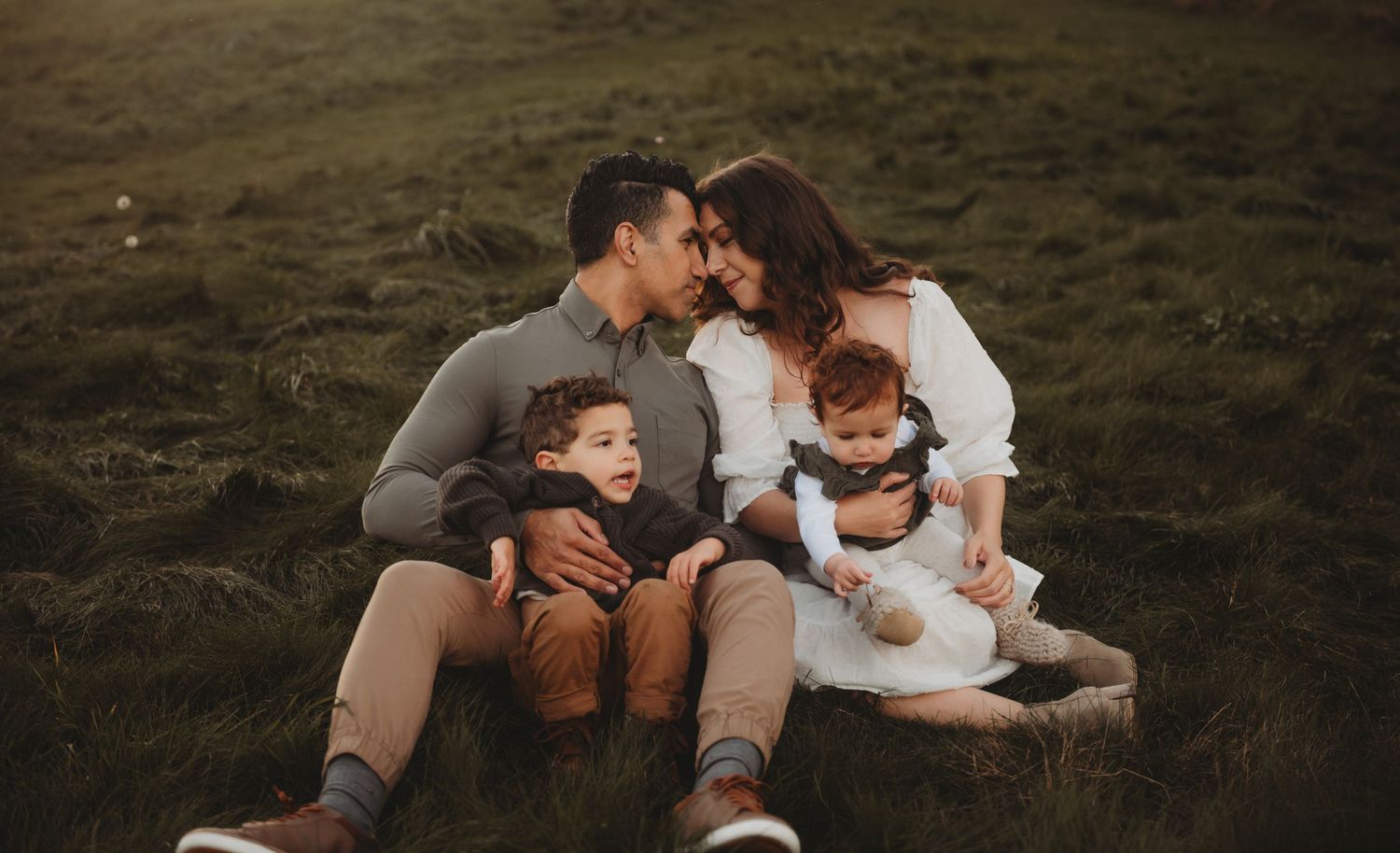 A loving family of four sits together on green grass, parents kissing while holding their two young children.