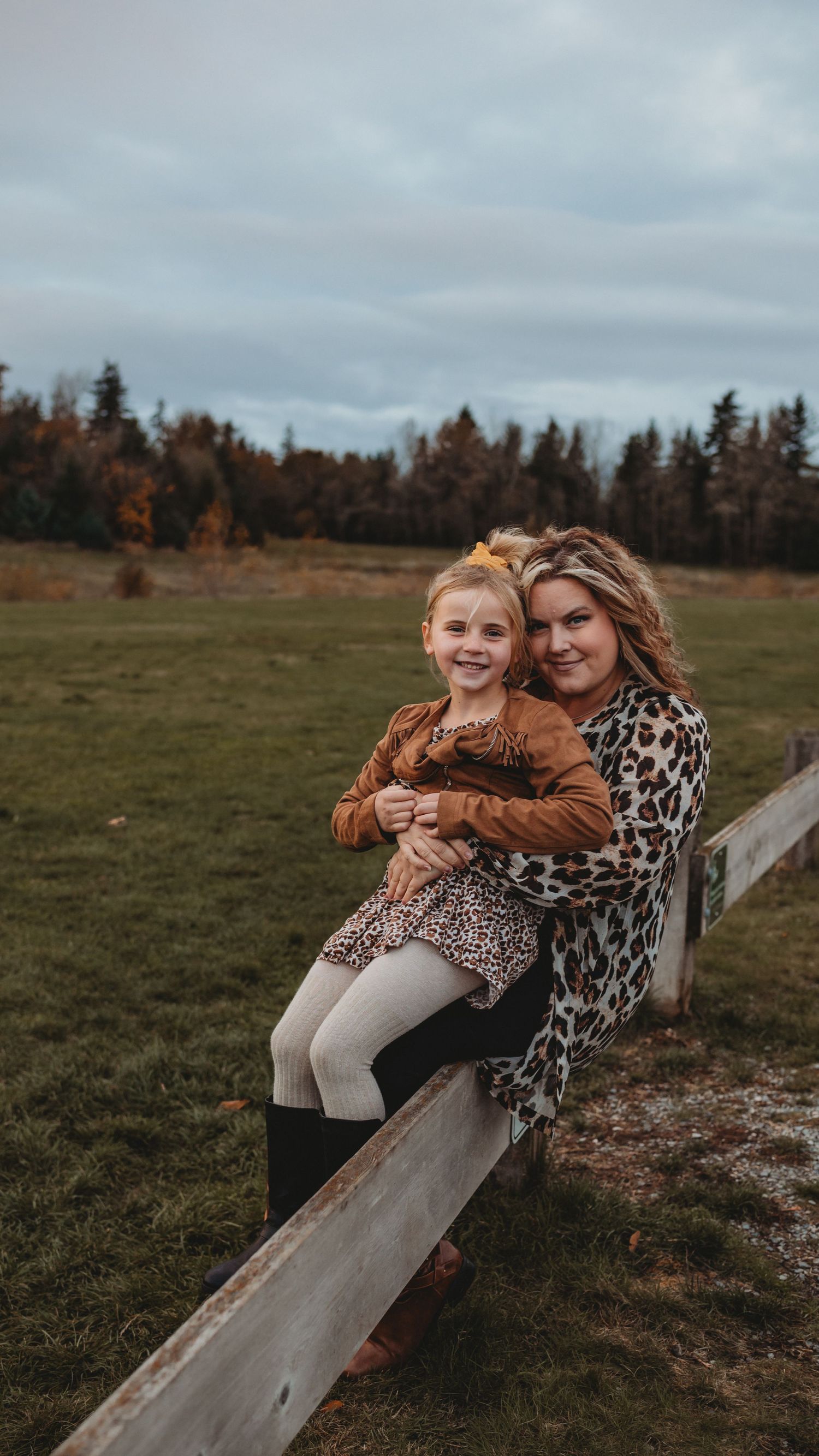 A smiling mother hugs her young blonde daughter while sitting on a fence in an autumn field.