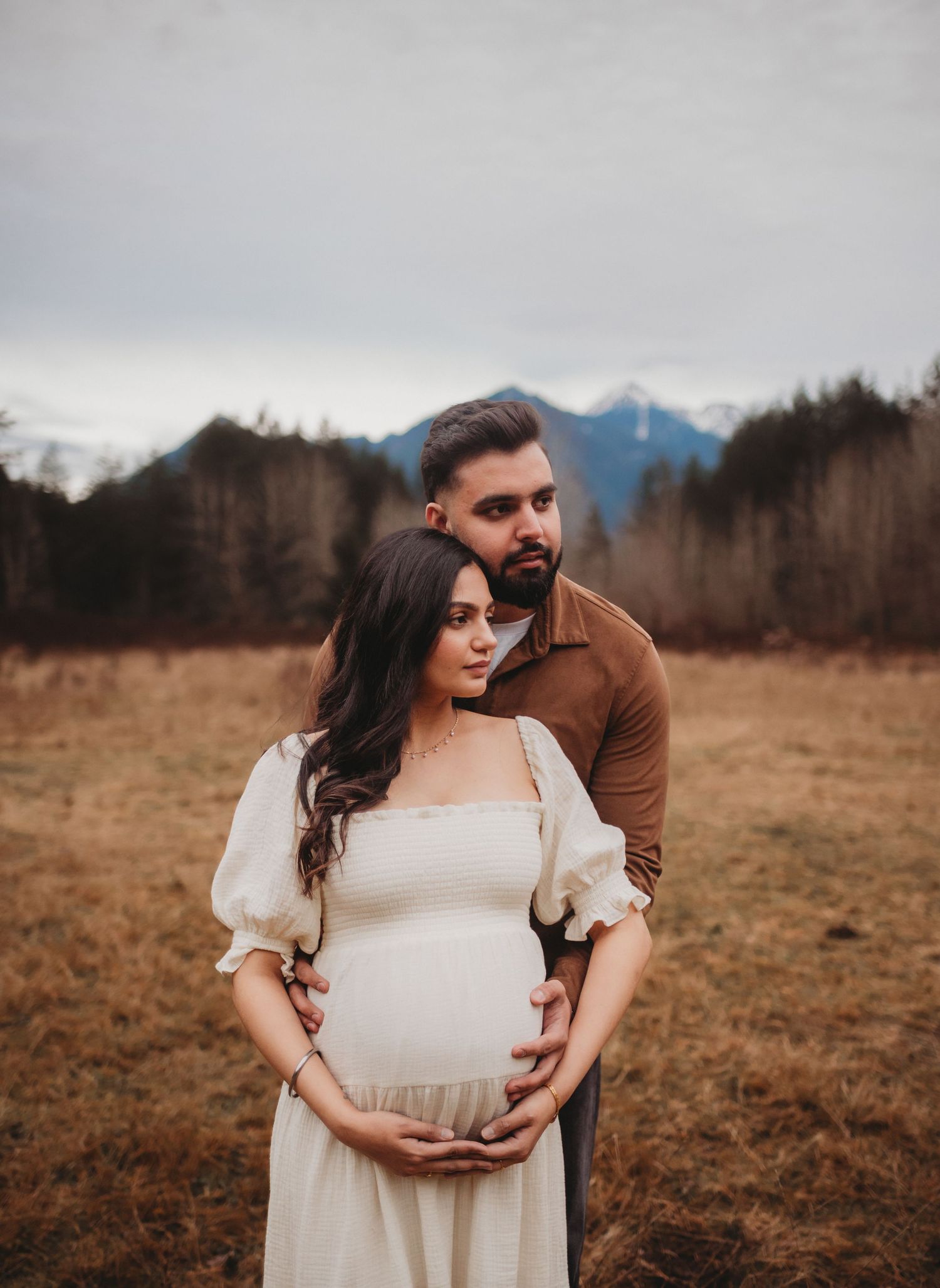 Man tenderly embraces pregnant woman in white dress during outdoor maternity shoot with mountain backdrop.