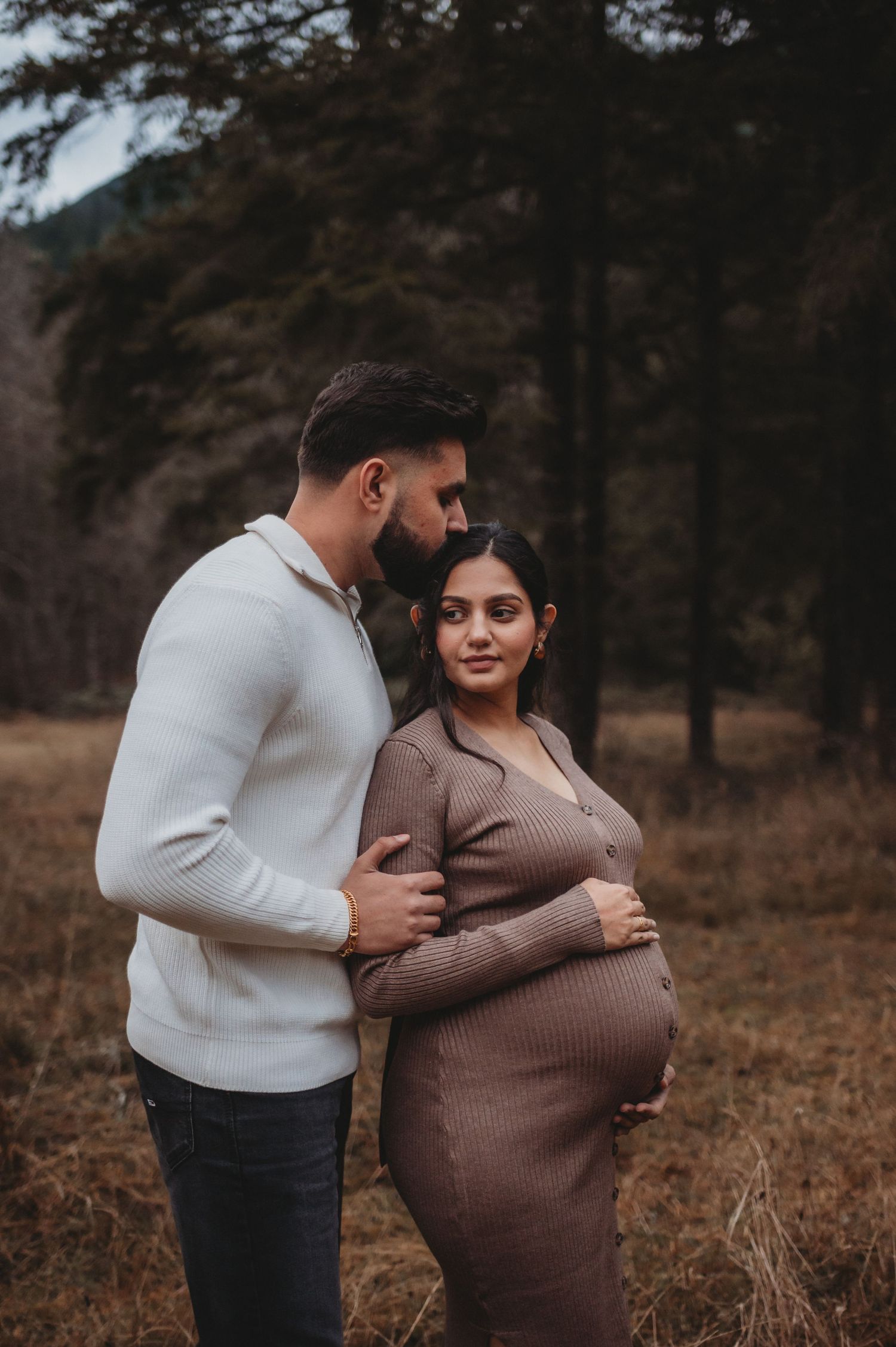 Man kissing pregnant woman's head while she smiles, standing in autumn forest maternity session.