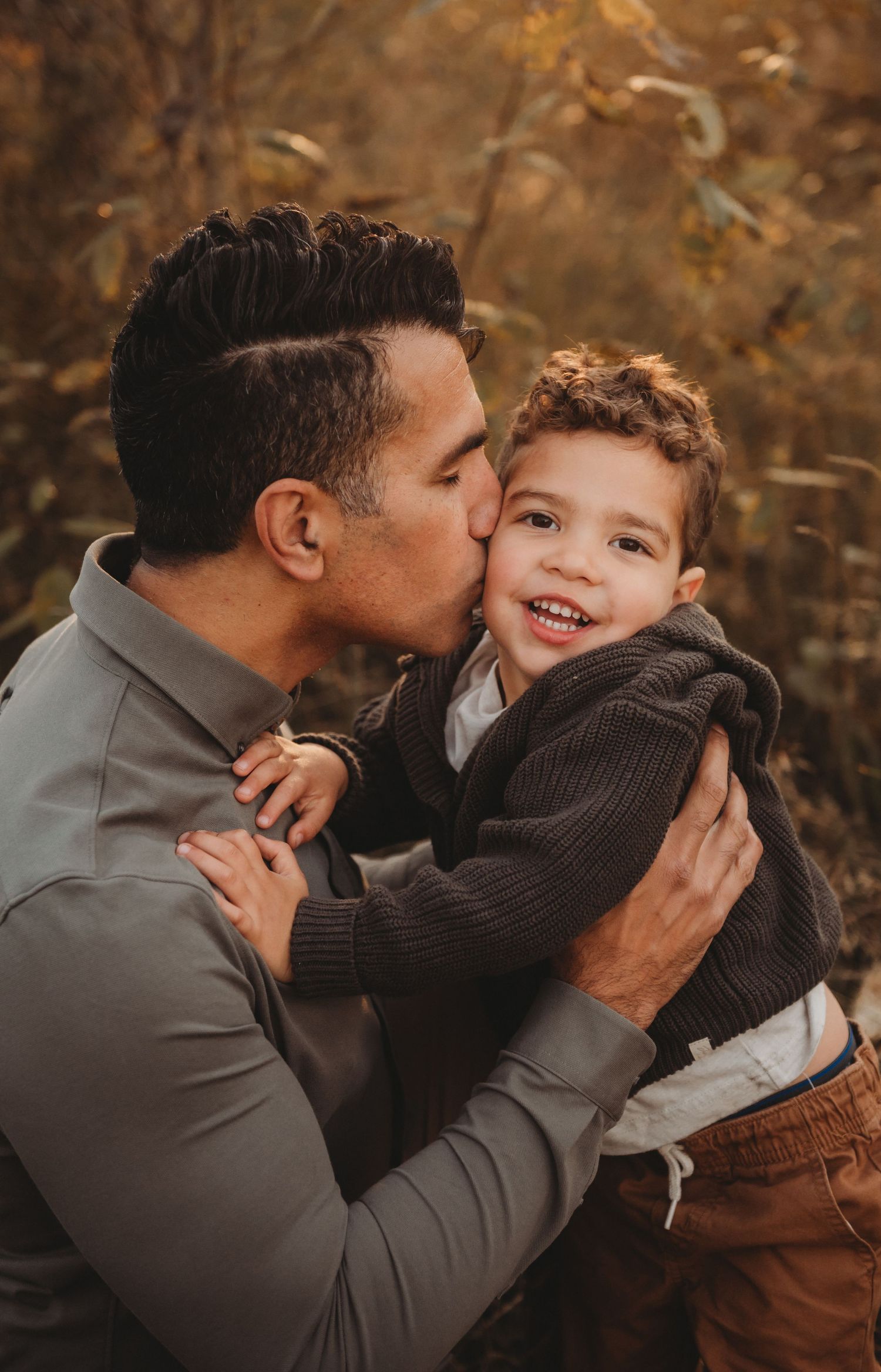 Father kissing his laughing young sons cheek while holding him in a golden autumn landscape.