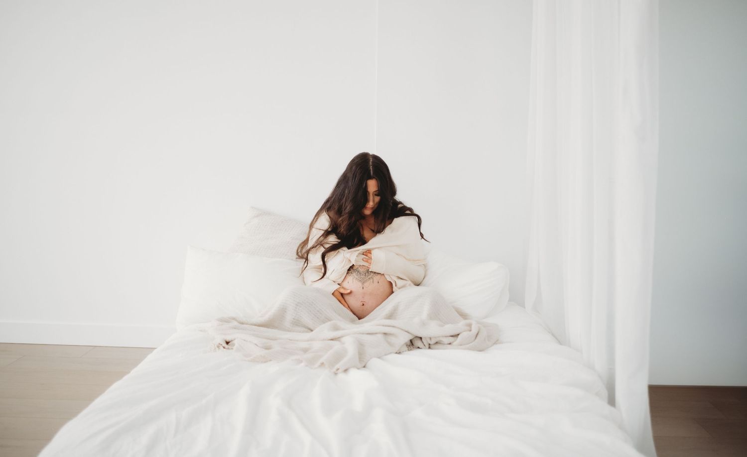A new mother sits on a white bed, tenderly cradling her newborn baby in soft natural light.