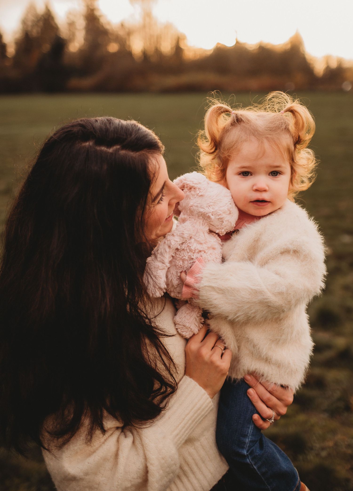 Dark-haired mother holding her curly-haired toddler girl with a pink stuffed animal.