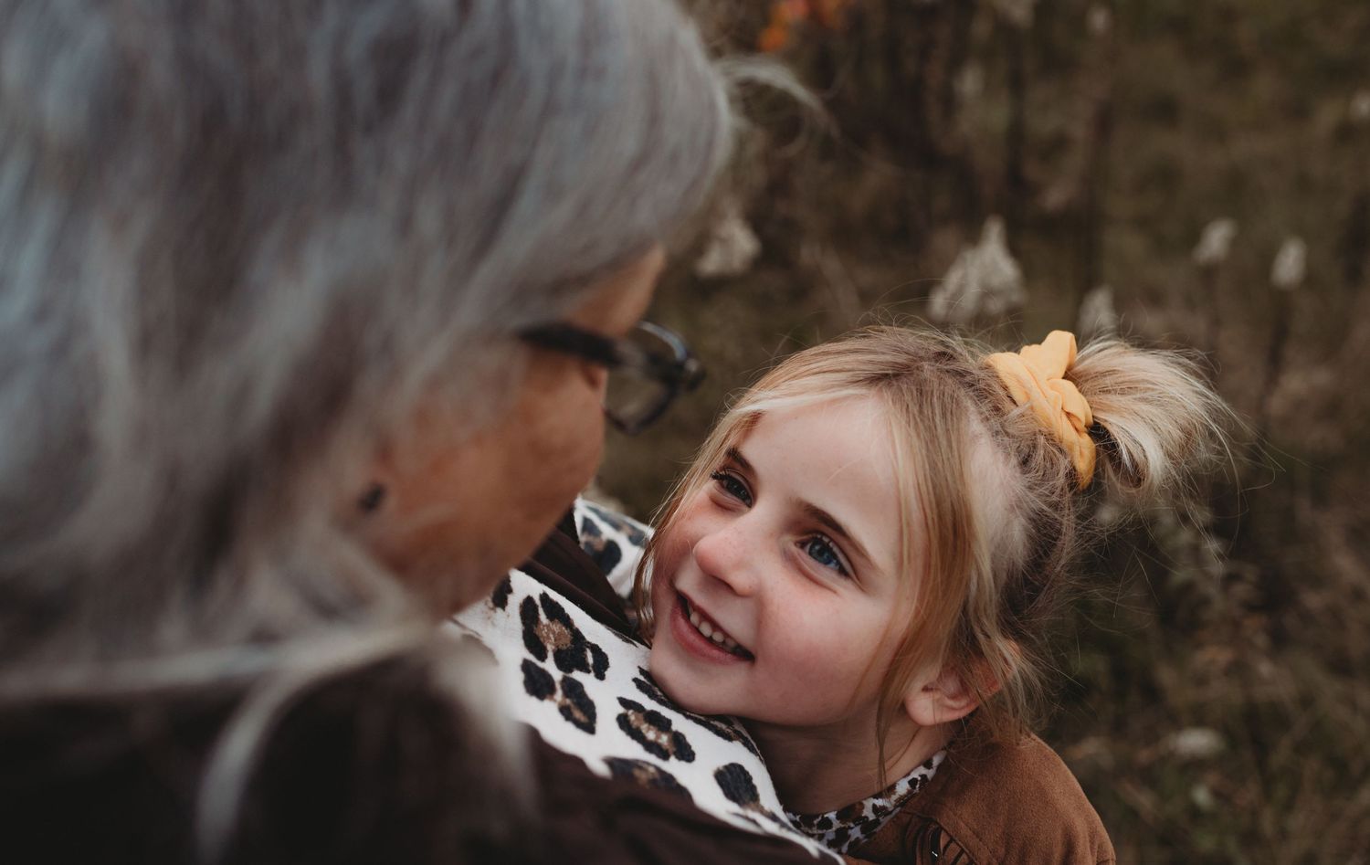 Young blonde girl with yellow bow smiling up at an older woman in autumn outdoors.