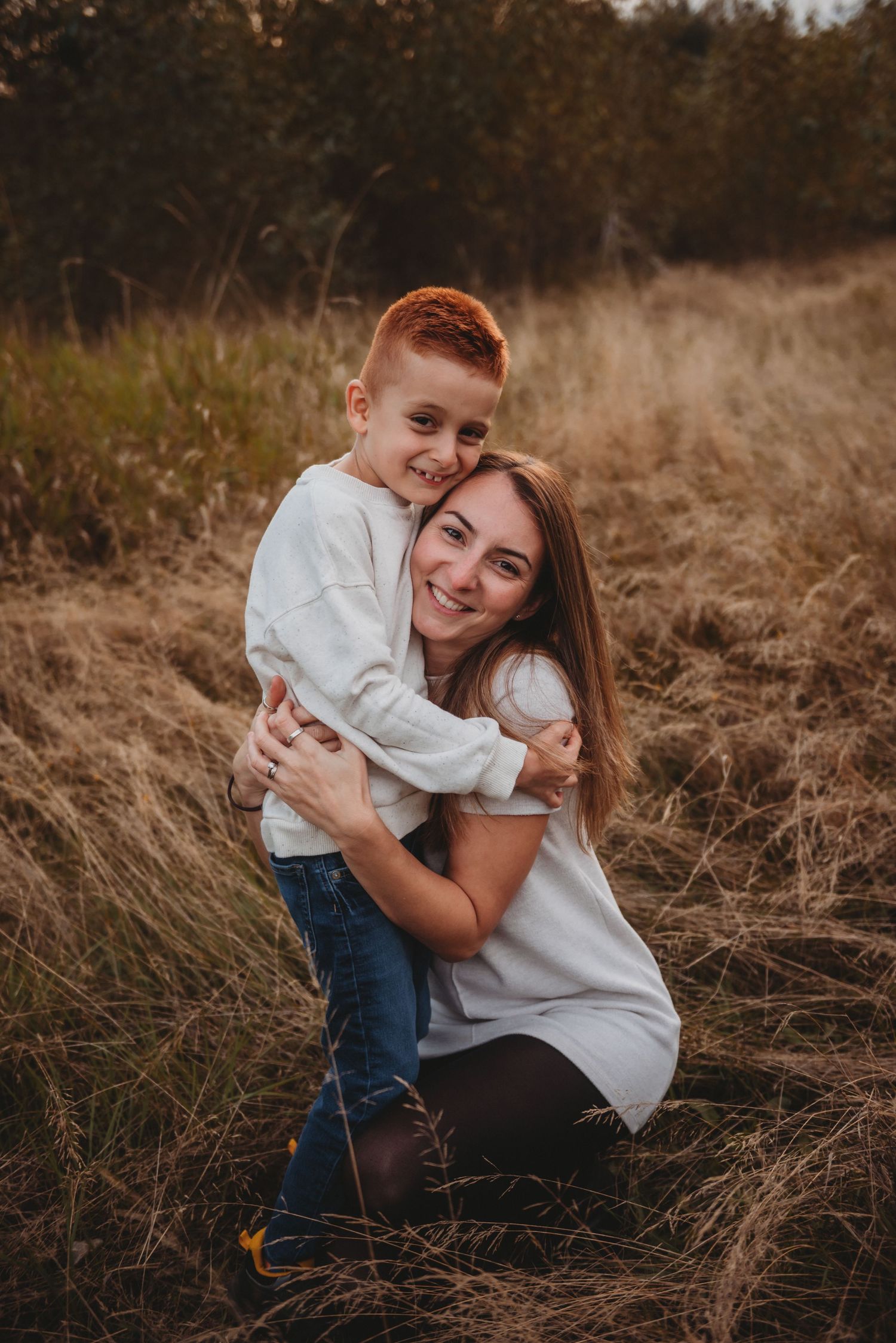 Joyful red-haired boy hugging smiling mother kneeling in golden autumn grass field.