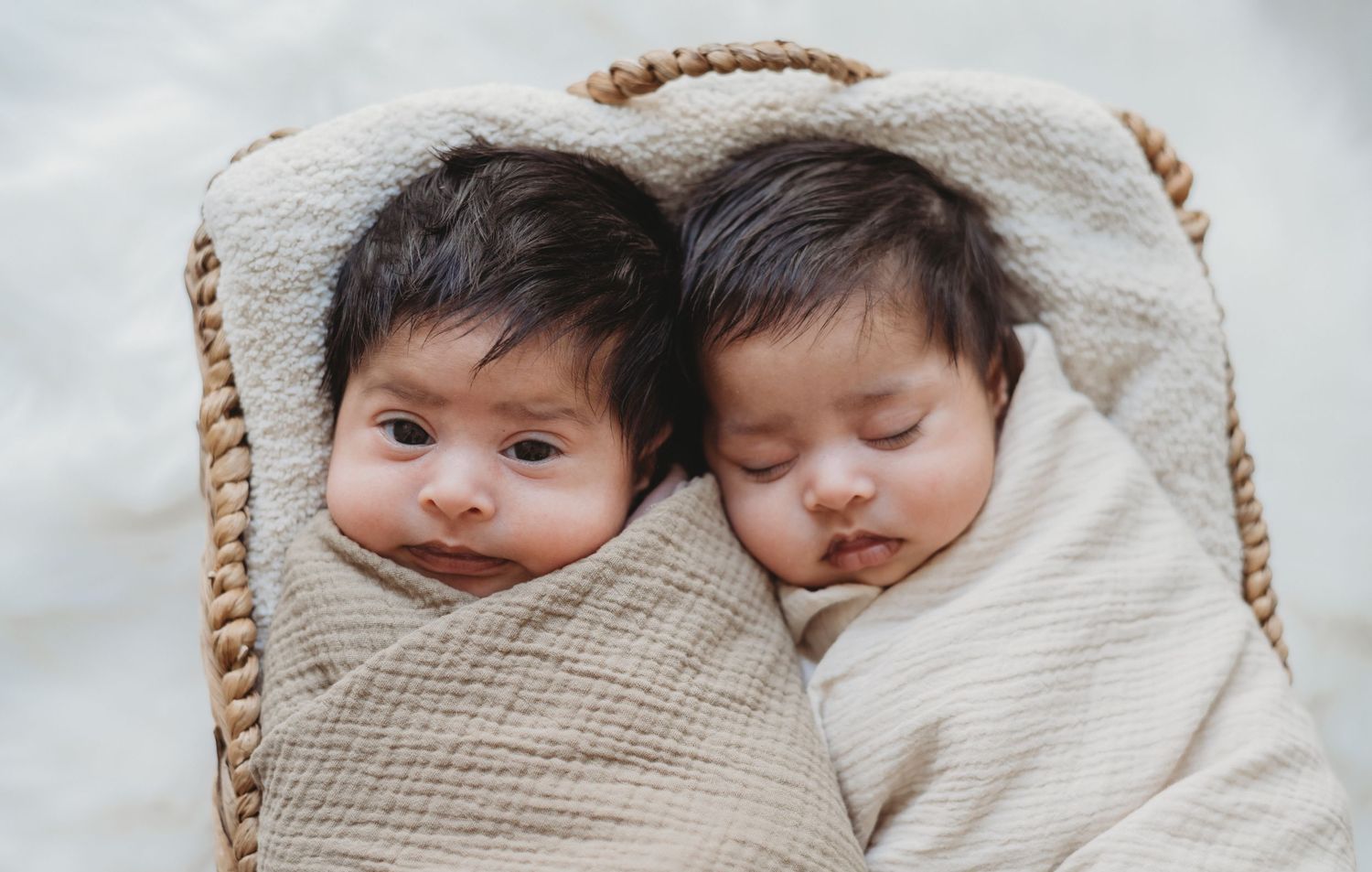 Two newborn twins swaddled together in cream muslin wrap inside cozy wicker basket.