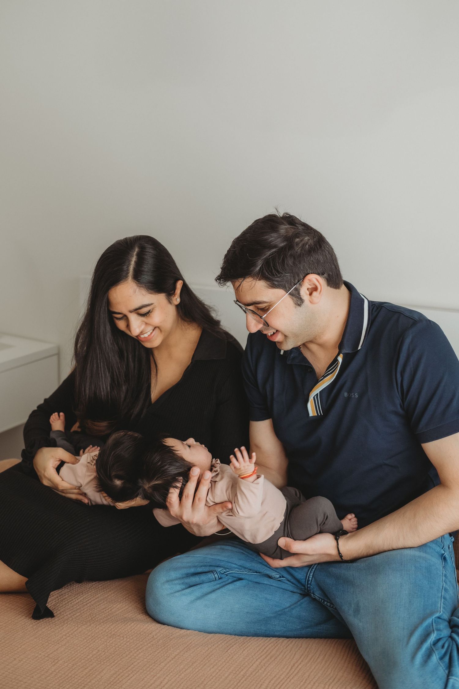 Happy parents sitting on floor smiling and holding their newborn twins dressed in pink outfits.