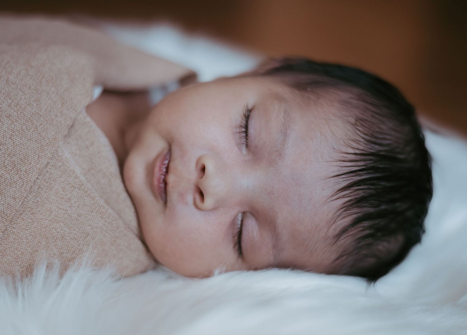 Peaceful newborn baby sleeping wrapped in a soft beige blanket on a white fluffy surface.