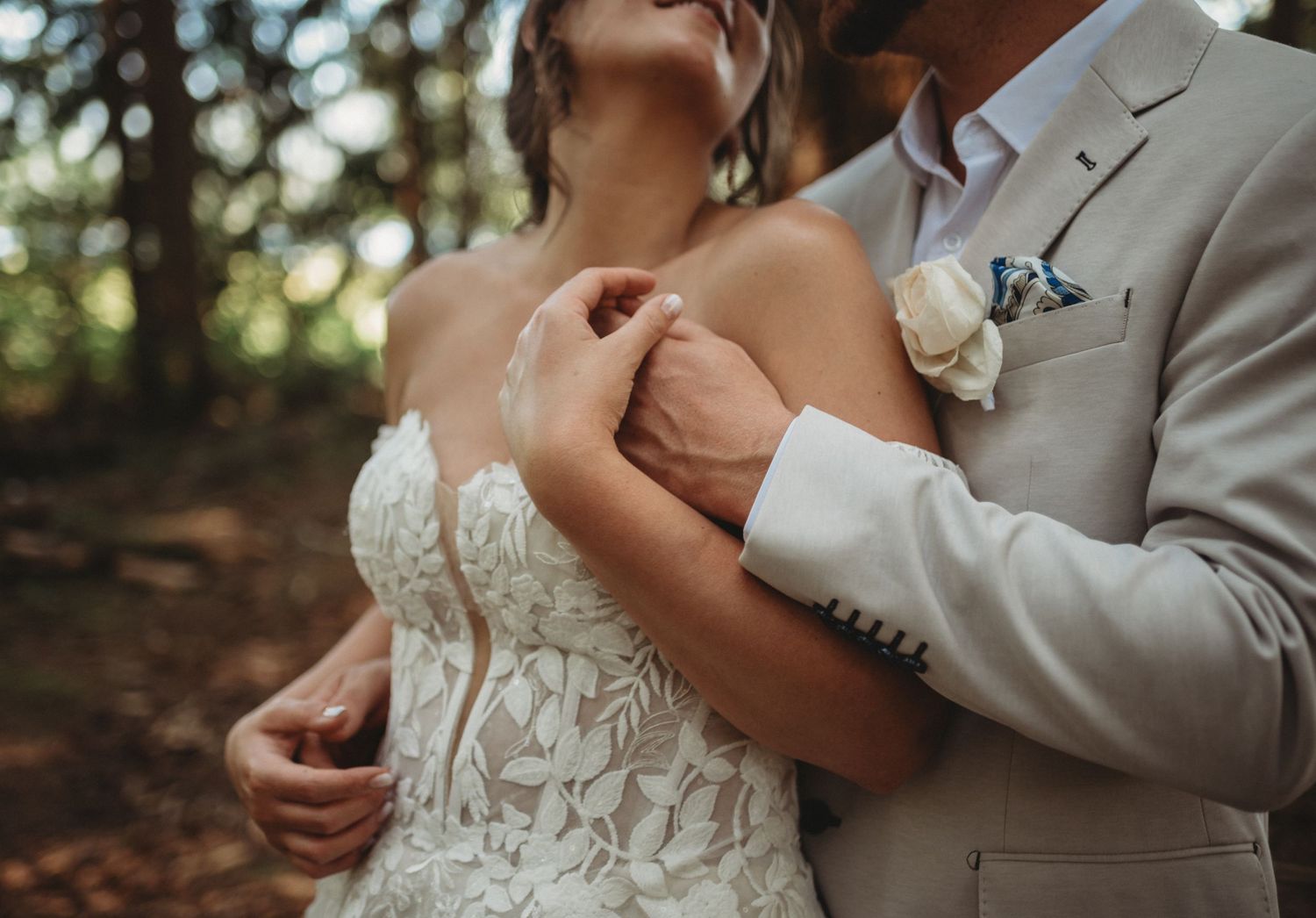 Groom in beige suit embraces bride in lace wedding dress in forest.