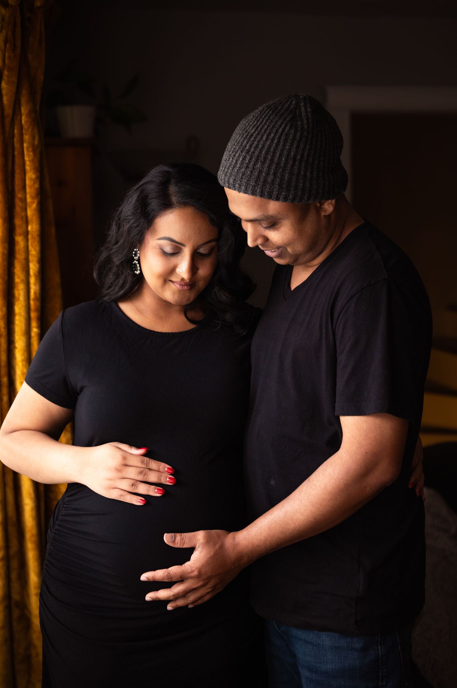 Pregnant couple in matching black outfits gently holding baby bump indoors with warm lighting.