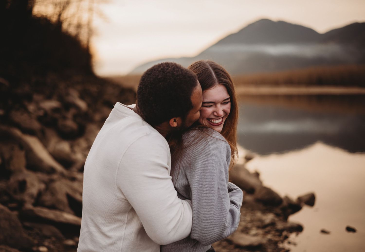 A man embraces a laughing woman by a mountain lake during a golden hour photo session.