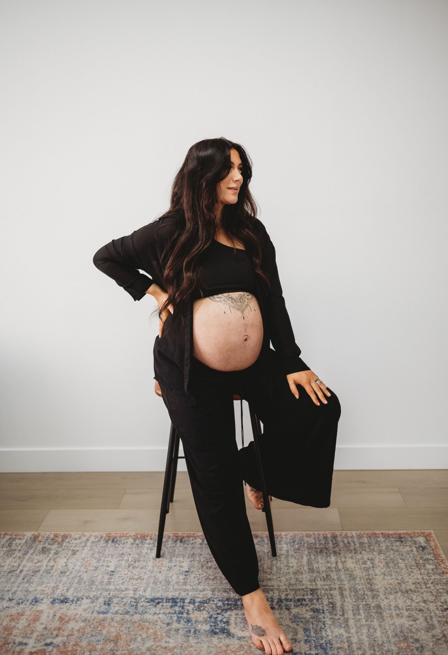 Pregnant woman in black outfit with tattoo showing baby bump beside chair.