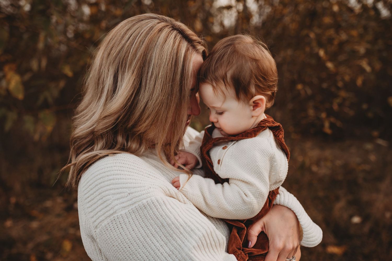 A mother tenderly holds her baby close in a warm autumn outdoor setting.