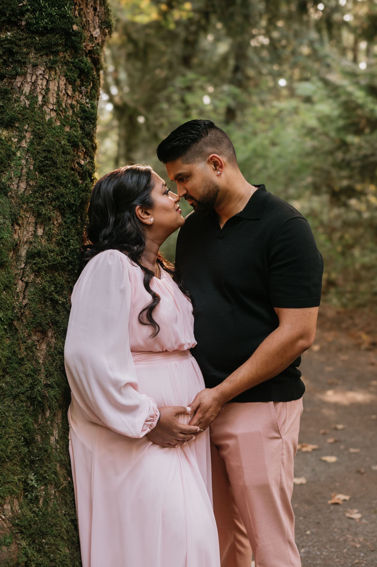 Expecting couple in pink and black outfits sharing intimate moment beside mossy forest tree.