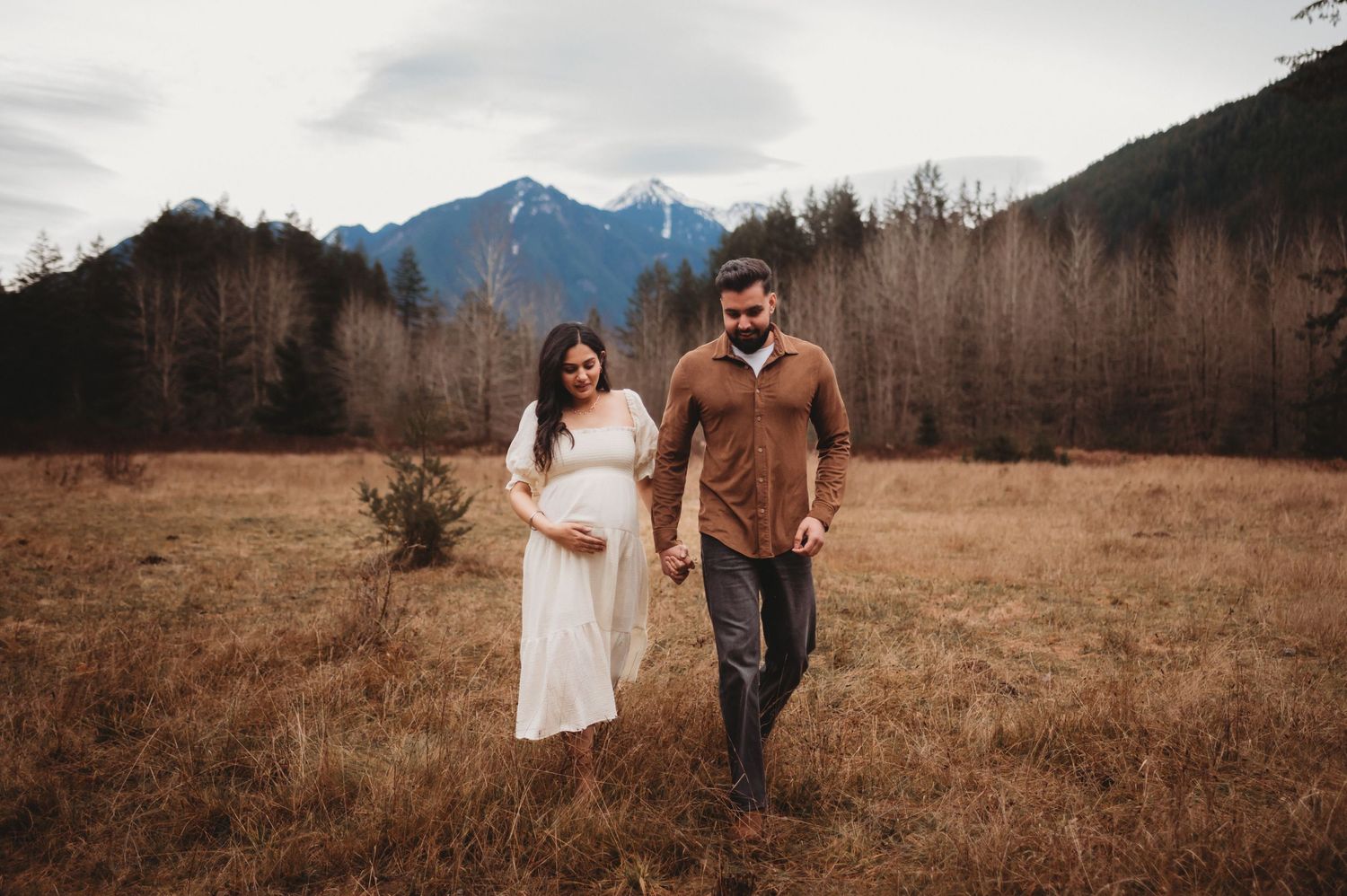 Pregnant couple strolling through rustic autumn field with majestic mountain range behind them.