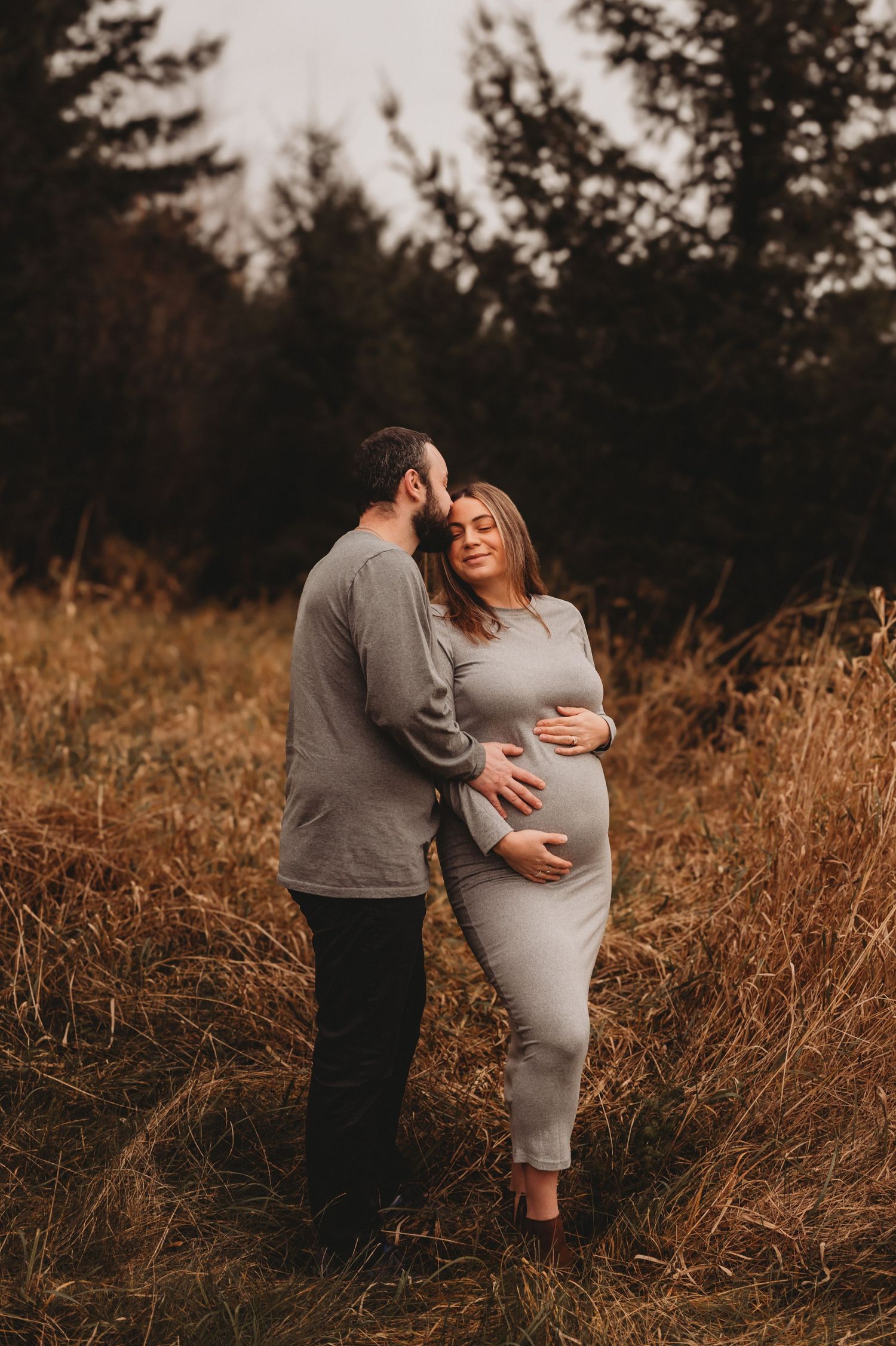 Bearded man kissing pregnant woman's head in golden grass field.