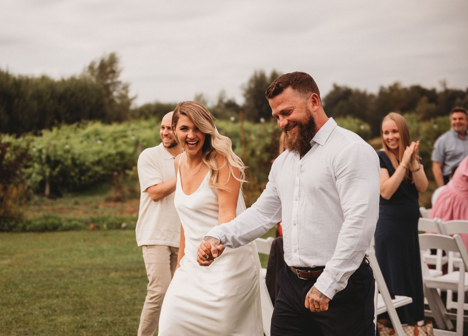 Smiling bride and groom walking hand in hand at outdoor wedding ceremony with guests applauding.