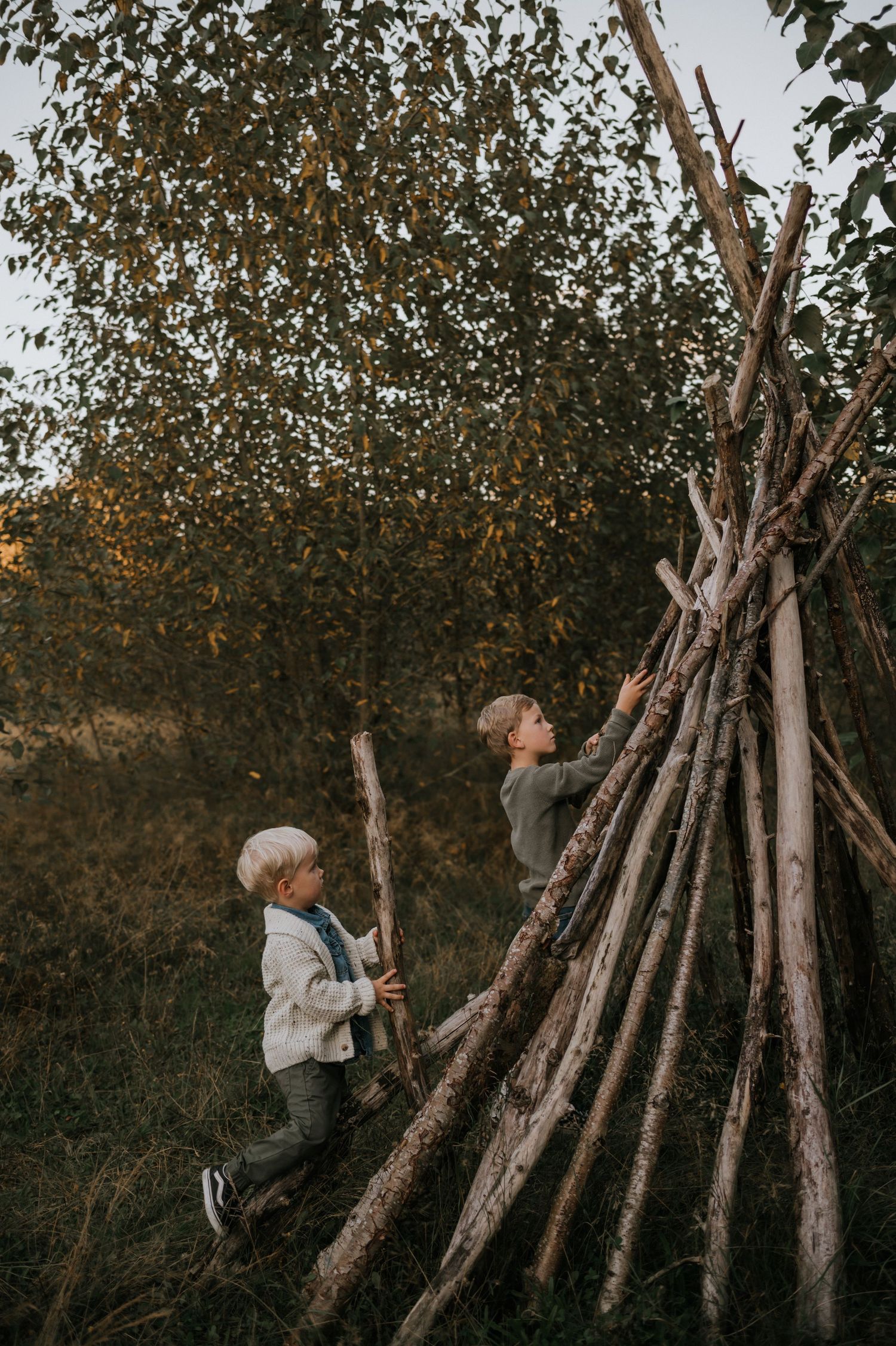 Two young boys playing and climbing a rustic wooden teepee structure in an autumn forest.