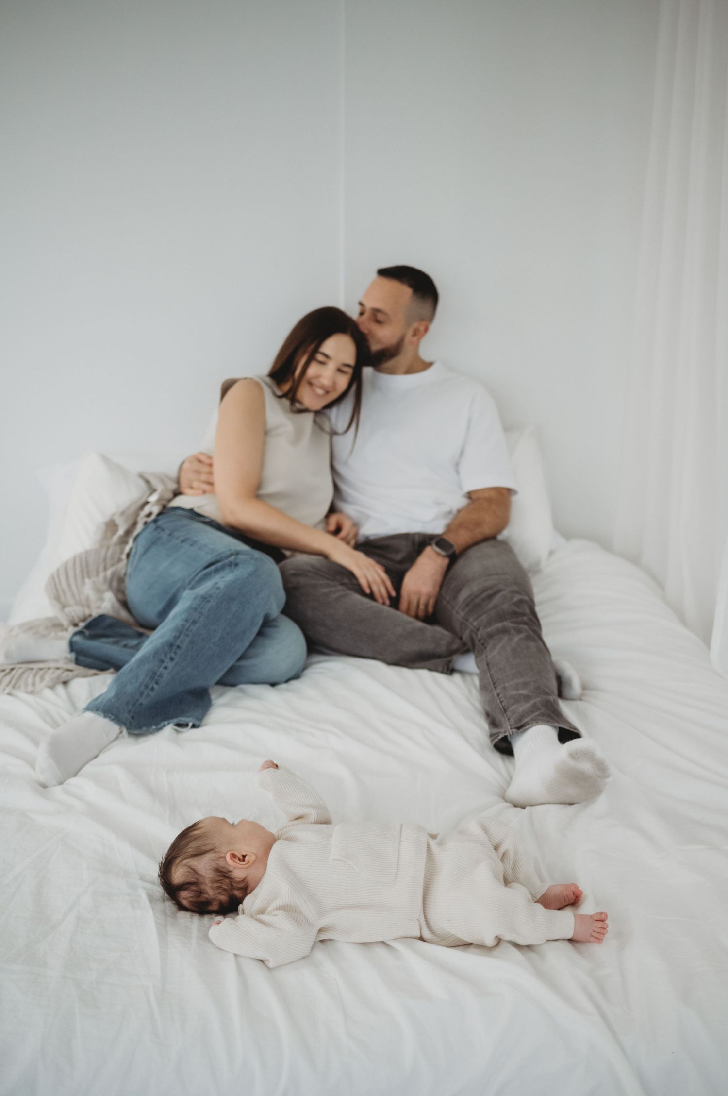 Young couple sitting on a white bed sharing a tender moment with their newborn baby.