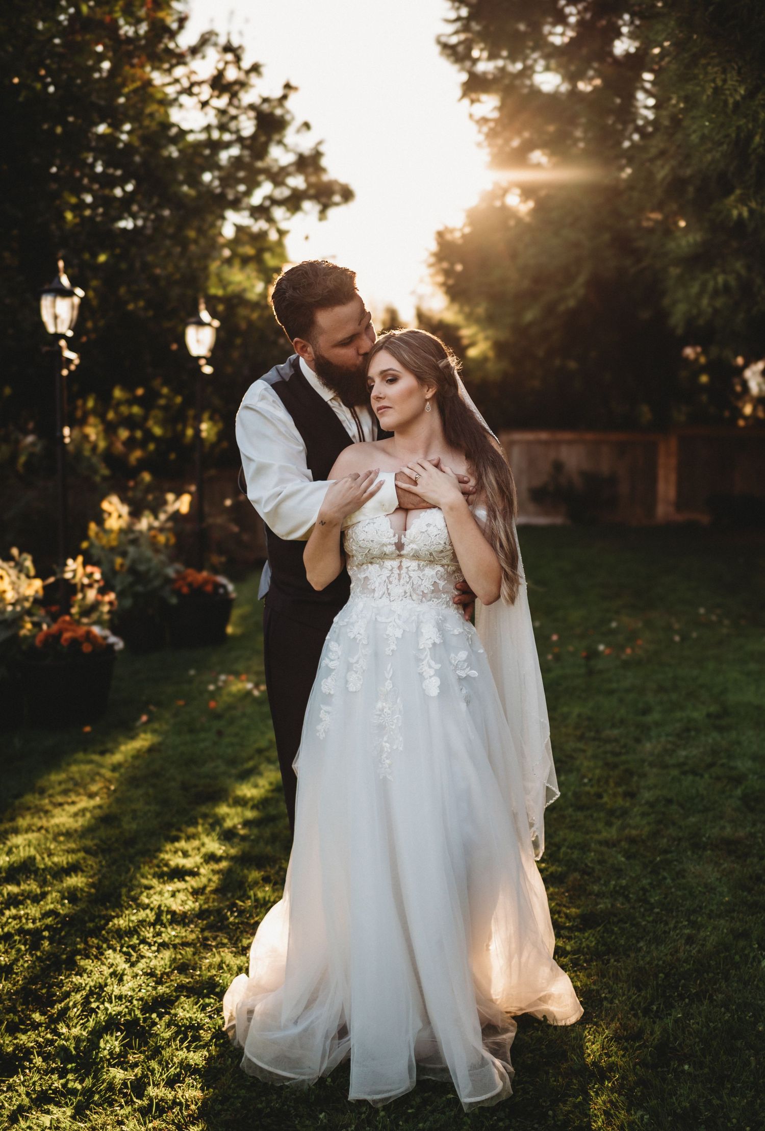 Groom standing behind bride in a stunning garden at golden hour, both posing in elegant wedding attire.