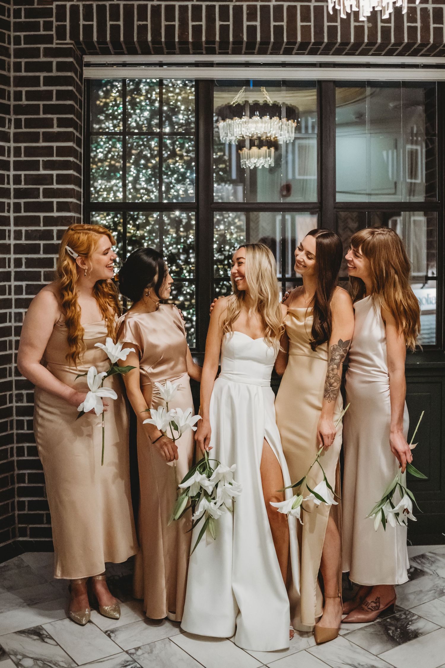 Bride in white gown smiles with bridesmaids in champagne dresses holding white lilies indoors.