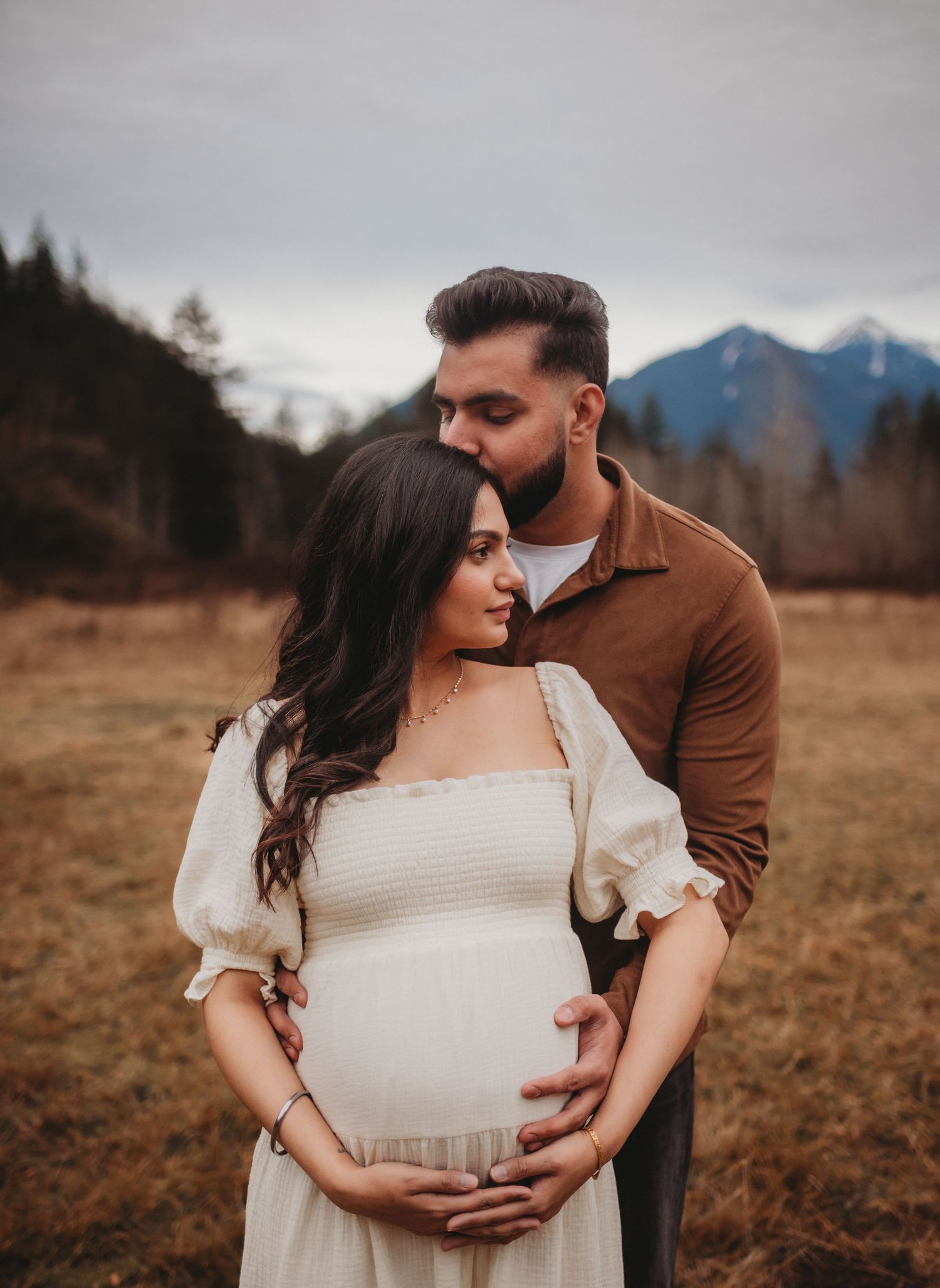 Man tenderly kissing pregnant partners head while cradling bump in mountain meadow.
