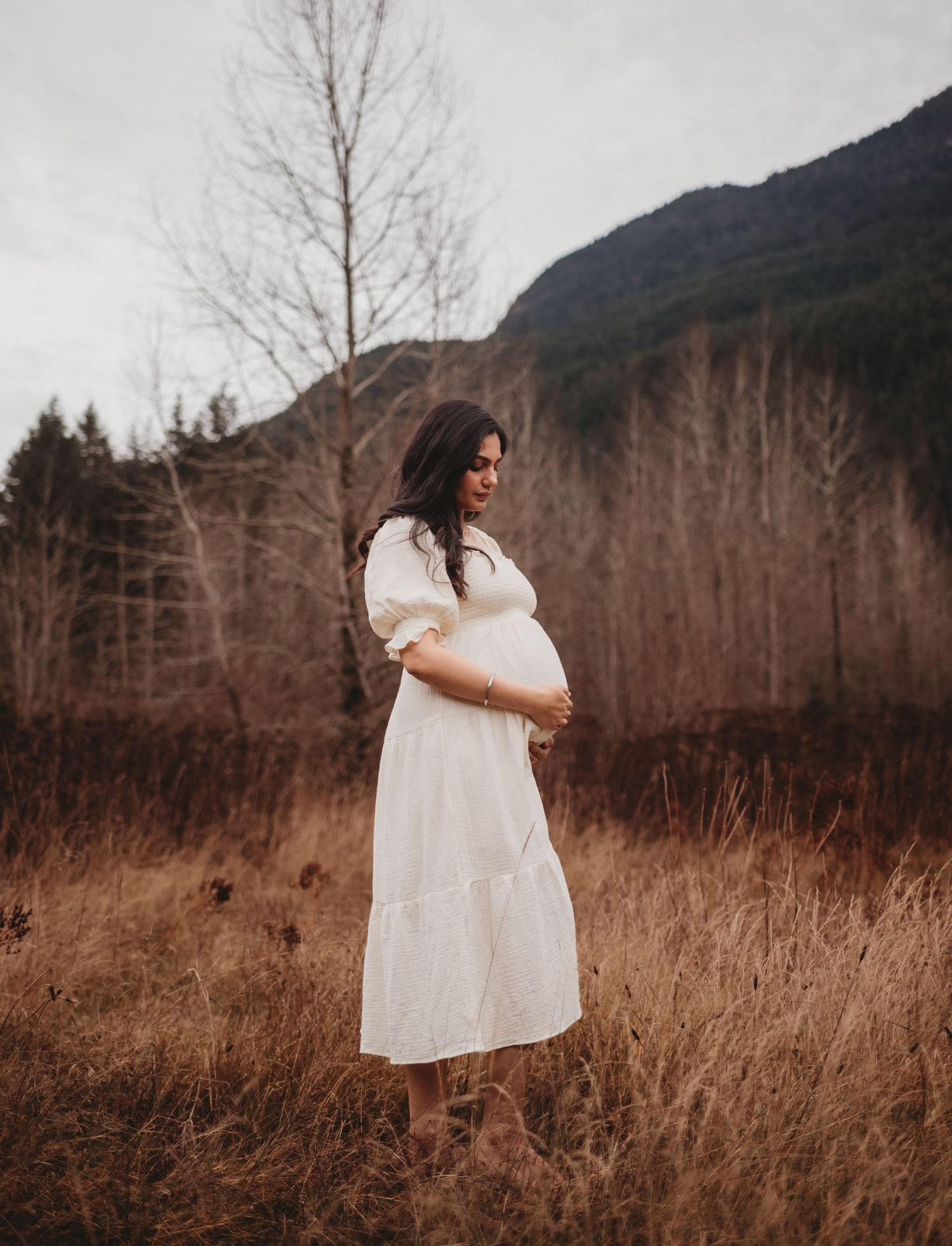 Expecting mother in white tiered dress cradling baby bump in autumn meadow with mountain backdrop.