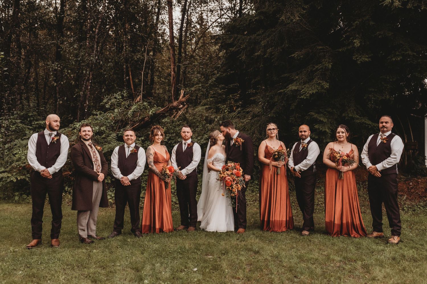 Wedding party poses outdoors in fall colors, bridesmaids in rust orange gowns and groomsmen in dark suits.