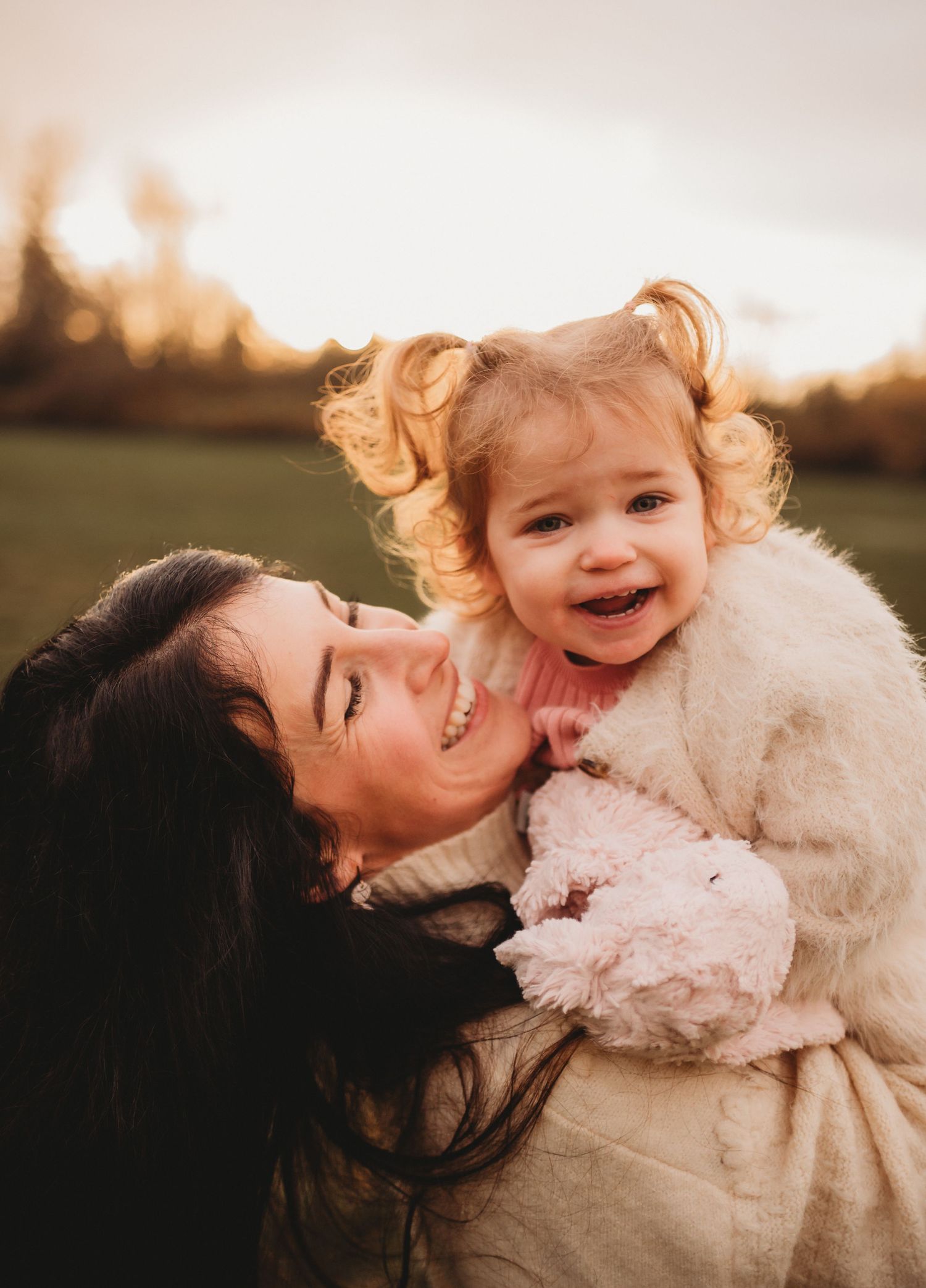 Smiling dark-haired mother cuddling her happy curly-haired toddler at golden hour.