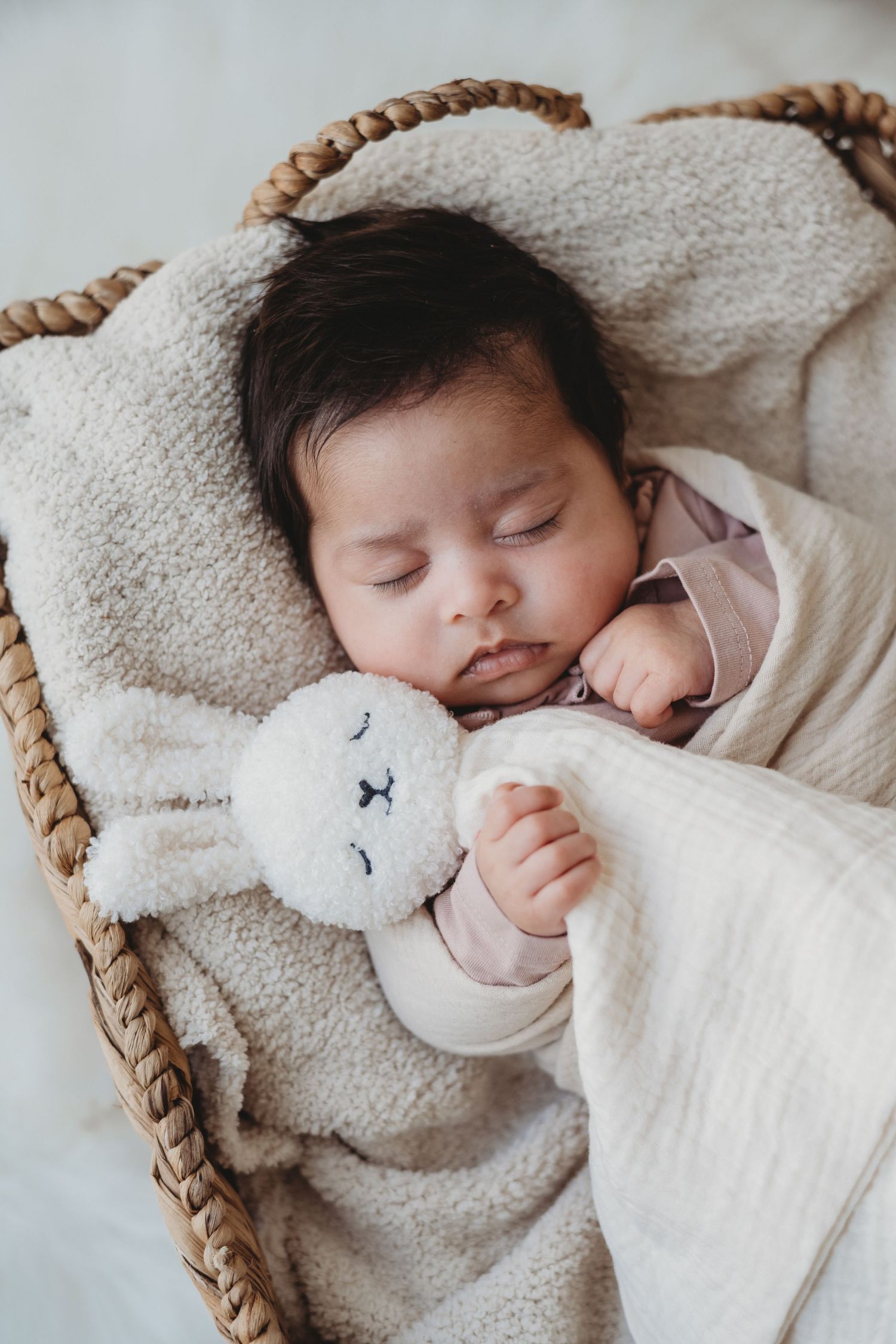 Newborn baby sleeping in wicker basket cuddling cream bunny stuffed animal lovey toy.