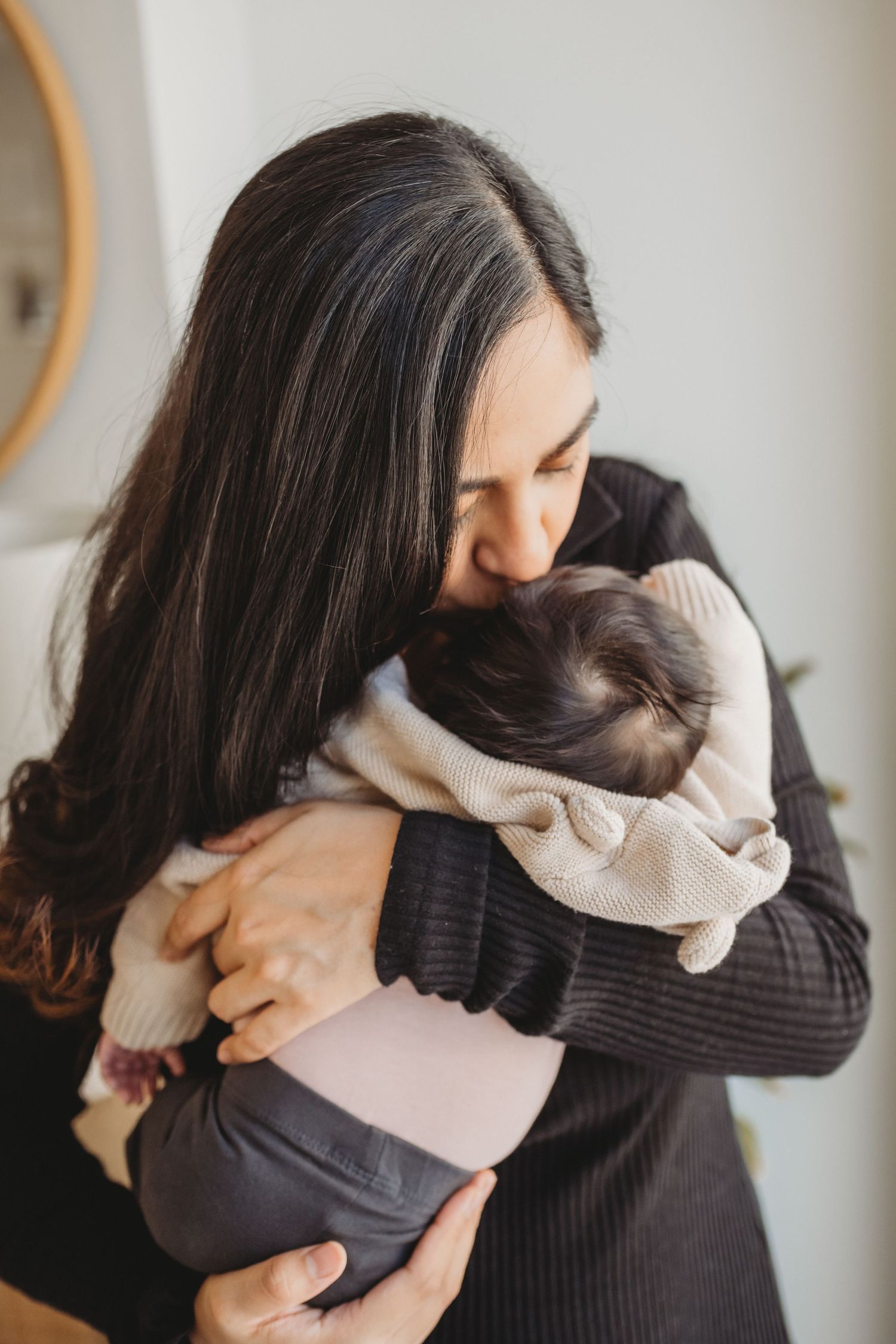 Mother tenderly embracing and kissing her newborn baby wrapped in a cream blanket indoors.