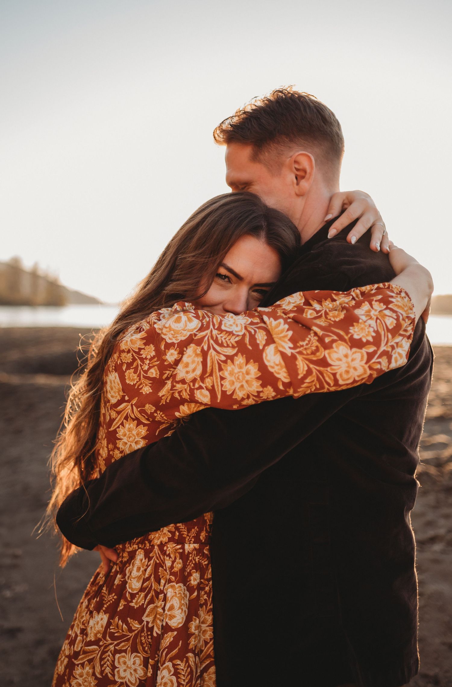 Woman in floral dress hugging man tightly on a sandy beach during warm golden hour light.