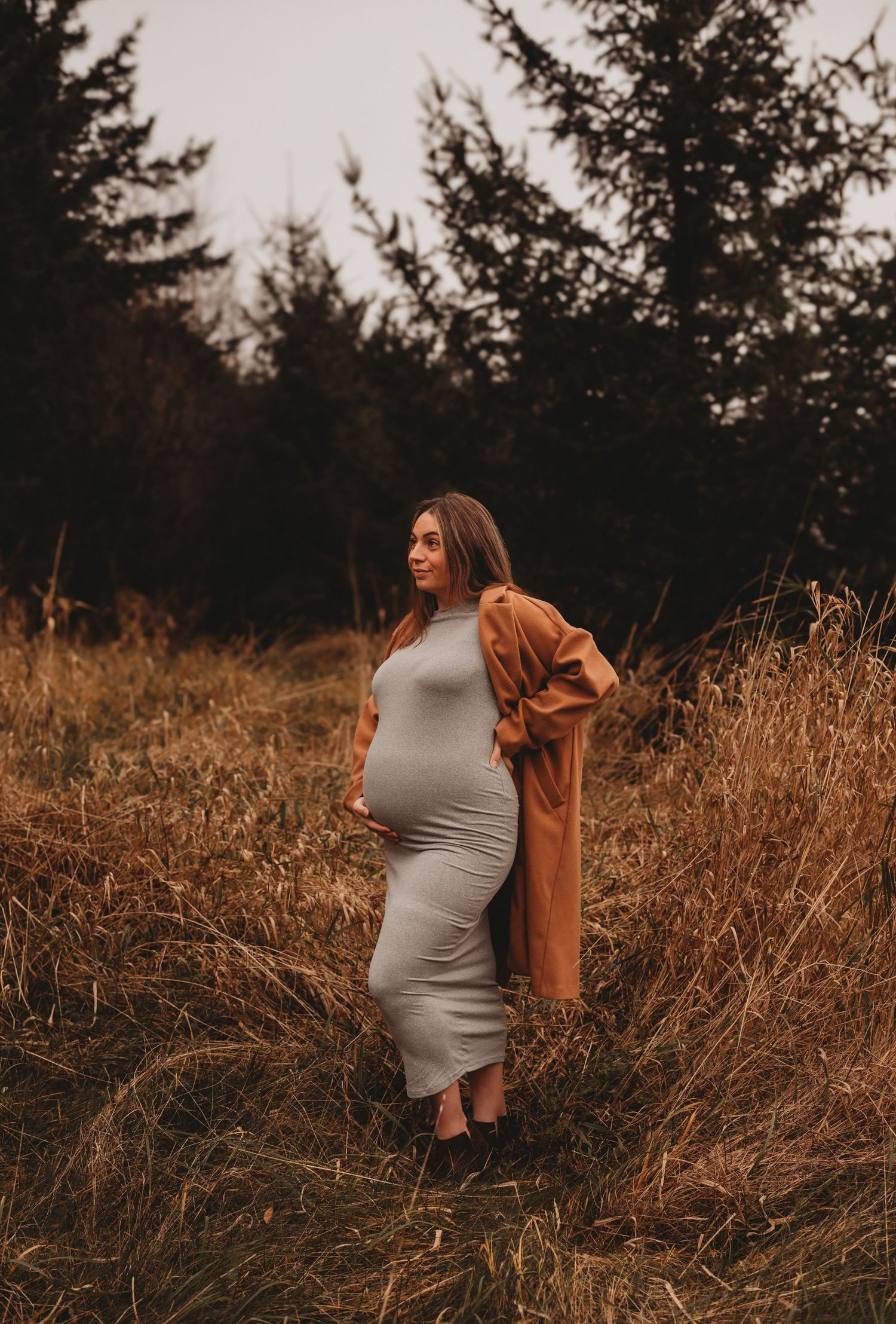 Pregnant woman in gray dress and camel coat standing in golden autumn field outdoors.