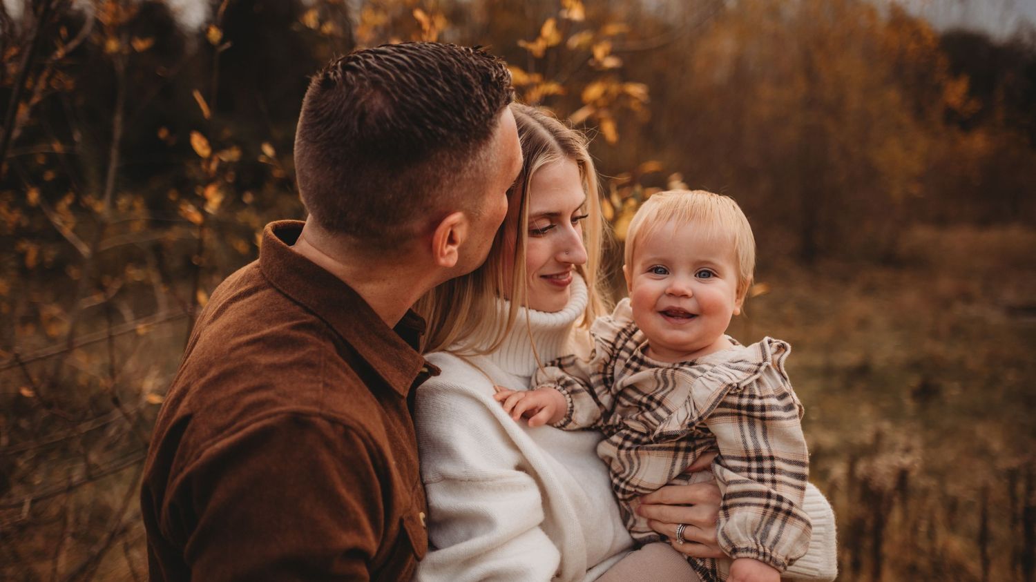 A couple smiles at their happy toddler during an autumn outdoor family photo session.