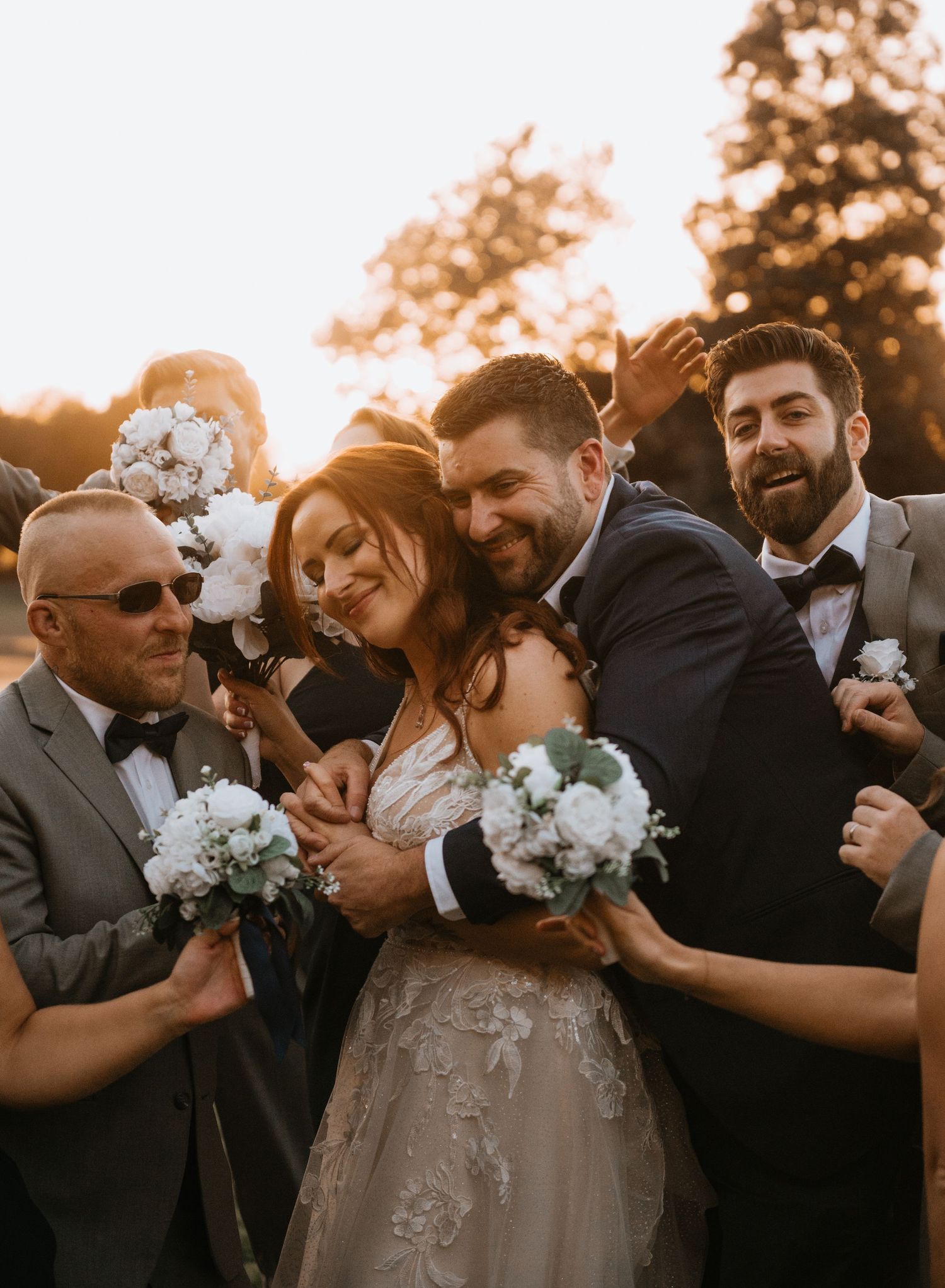 Joyful wedding party surrounds bride and groom in a warm golden sunset group hug.