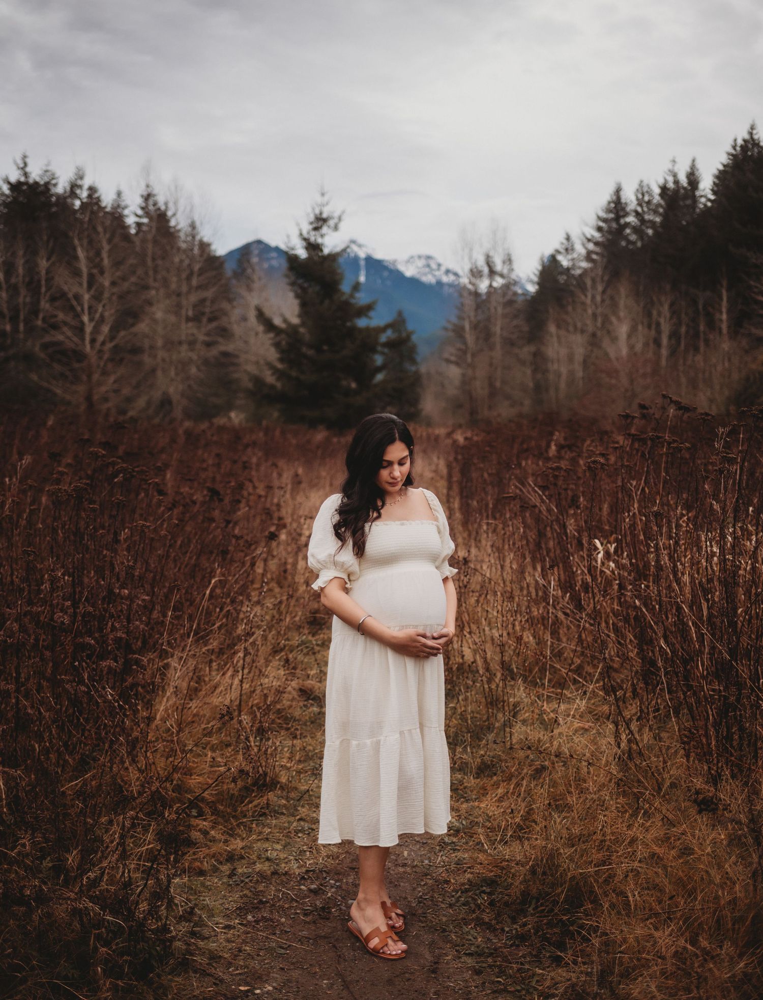 Pregnant woman in white dress stands among tall golden grasses with mountain forest background.
