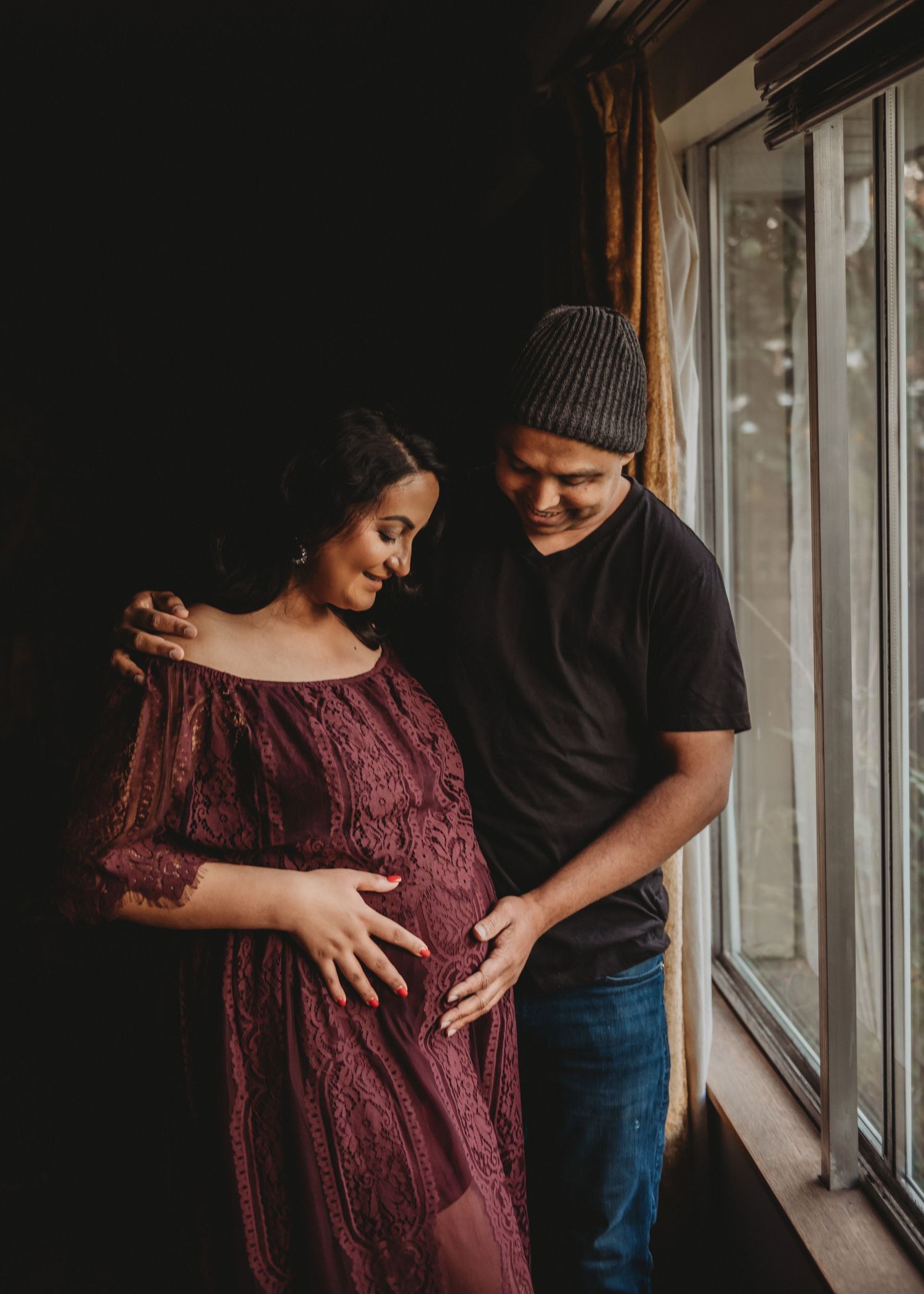 Expecting couple in burgundy lace dress and black outfit tenderly holding baby bump by window.